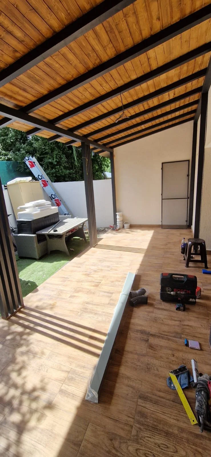 Under construction patio area with wooden flooring, partially installed white railing, tools, and construction materials. Outdoor seating with stacked tiles and insulation in background.