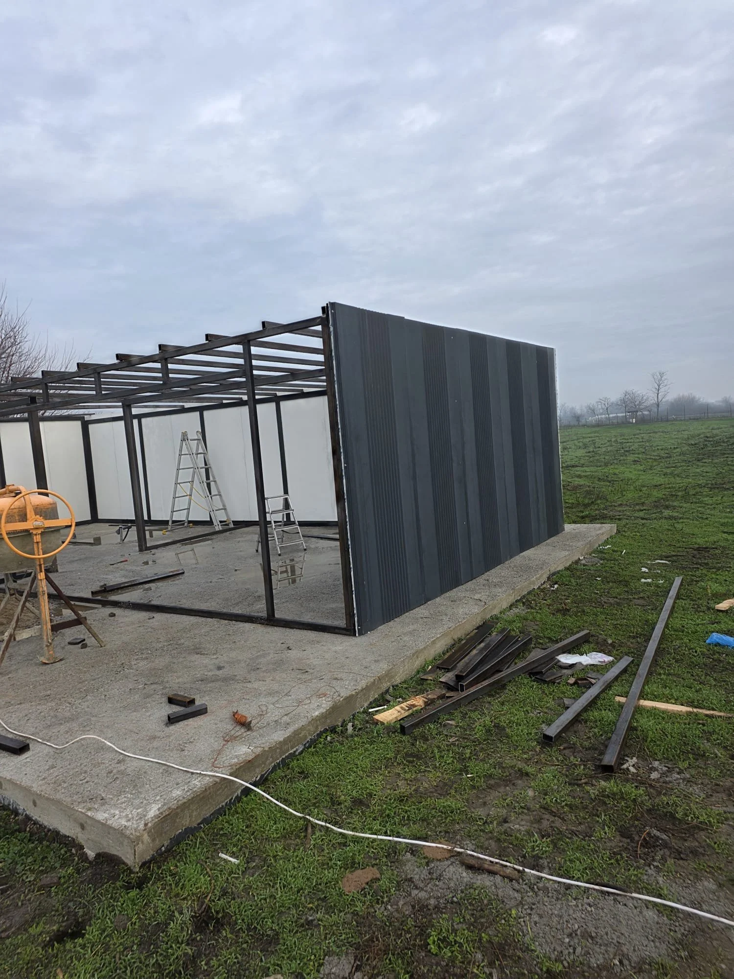 Construction site with a partially built metal structure on a concrete foundation, metal panels on the right side, ladders, and scattered building materials, with an overcast sky and open field in the background.