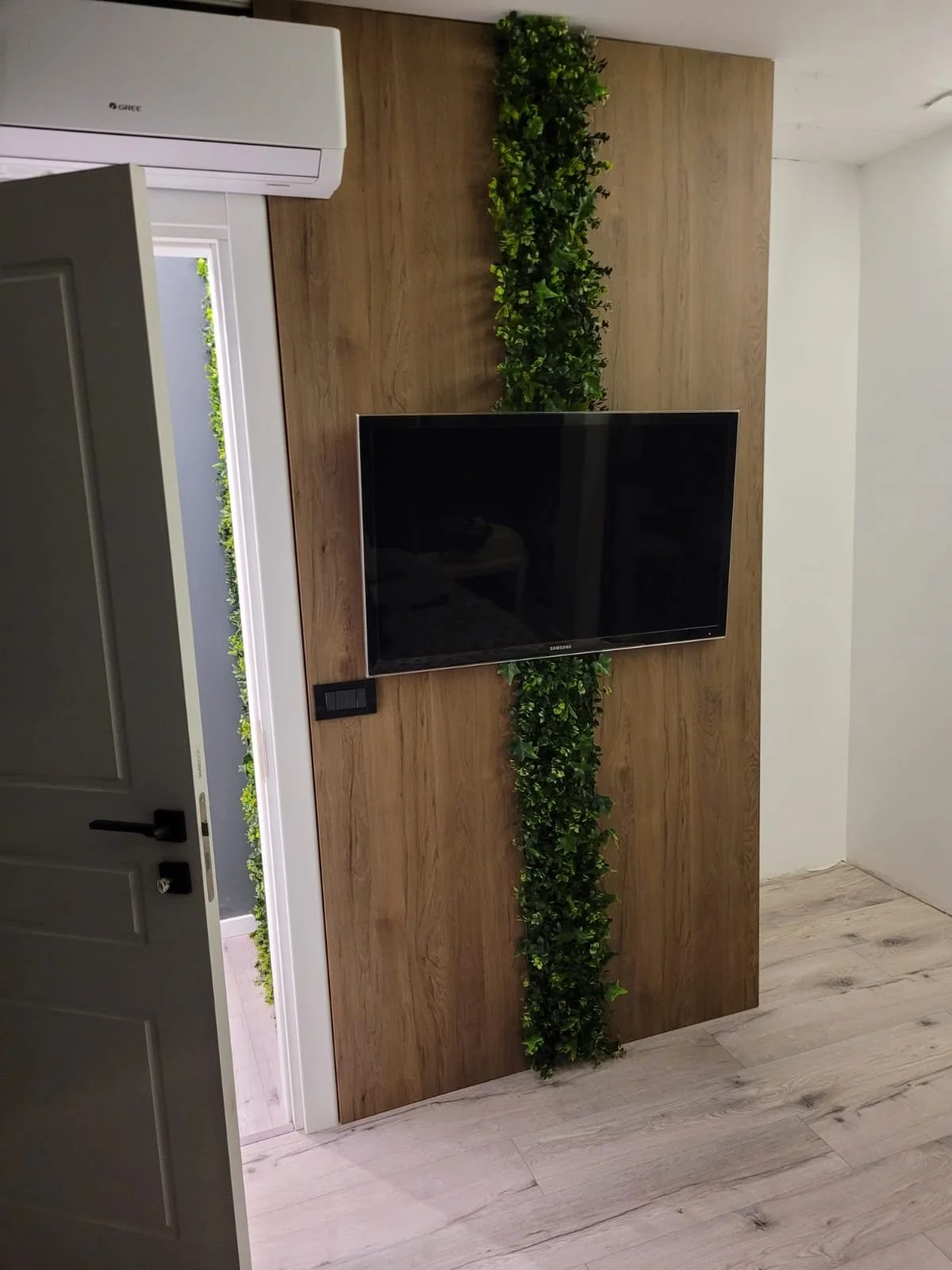 Living room with a wall-mounted TV on a wooden accent wall that has a green plant strip running vertically behind the TV, a door slightly open to an outdoor space with greenery, and an air conditioning unit above the door.