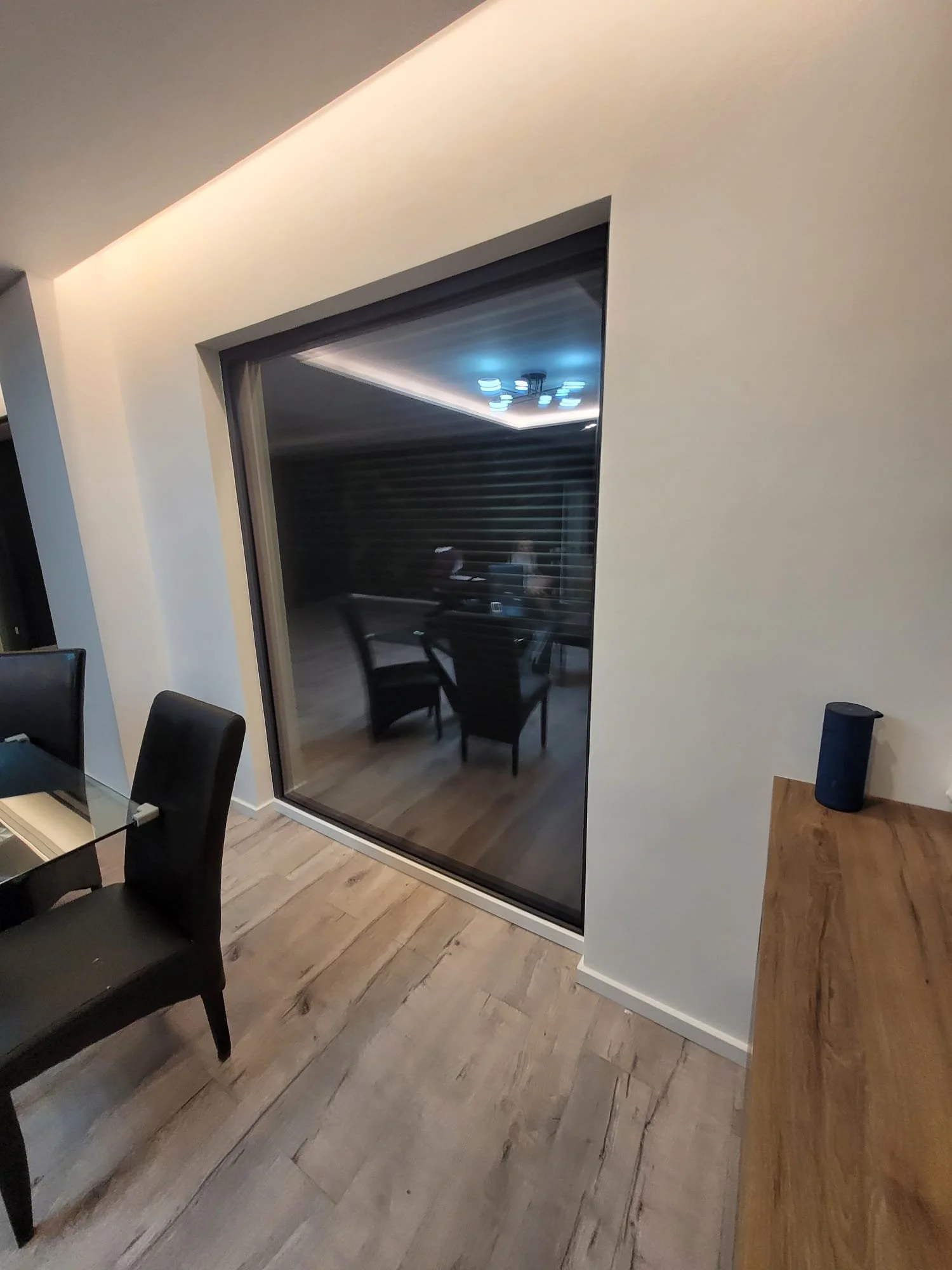Modern dining room with a large glass sliding door leading to an outdoor patio with table and chairs, hardwood floors, and a white wall with a wooden side table and blue speaker.