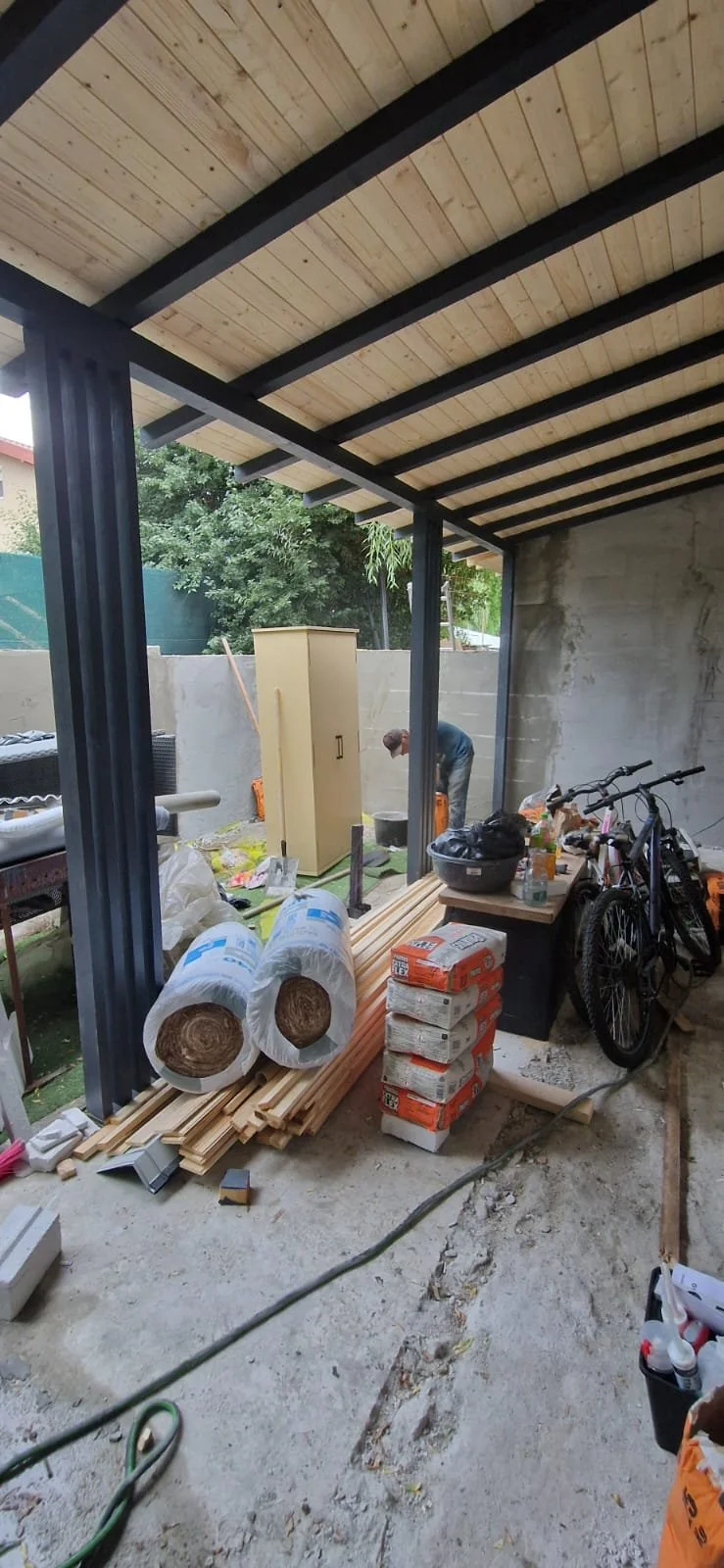 A covered outdoor area with construction materials including wood, insulation rolls, and boxes, with tools and two bicycles nearby, and a worker in the background working near a wall.