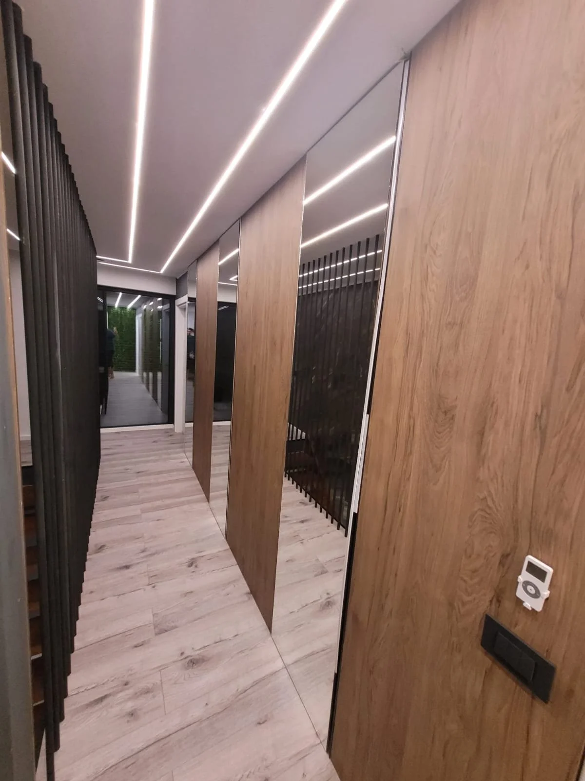 Modern hallway with wood-paneled walls, black metal accents, and sleek ceiling lighting, leading to a door with greenery visible outside.