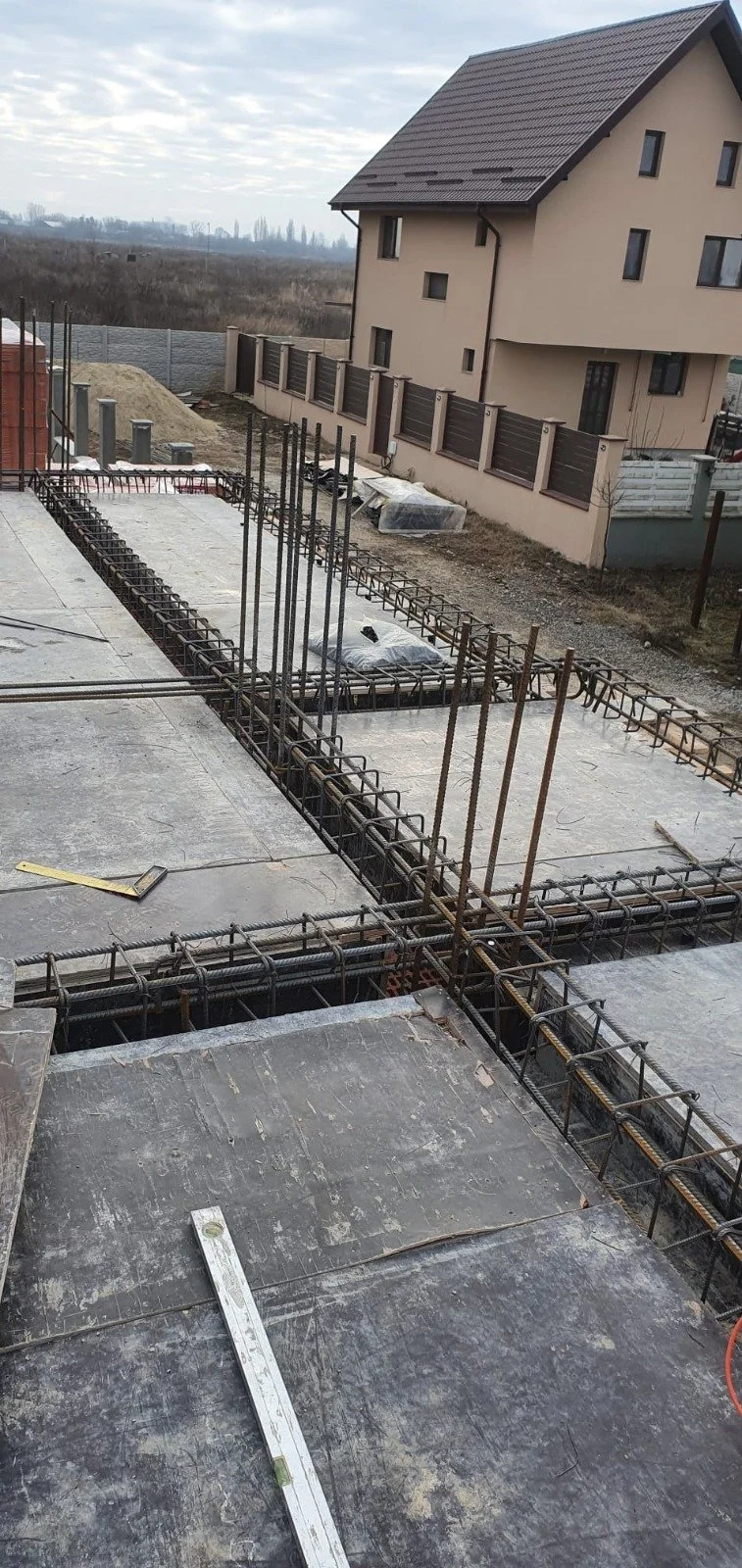 Construction site with rebar framework for concrete foundation, adjacent to a residential house, on cloudy day.