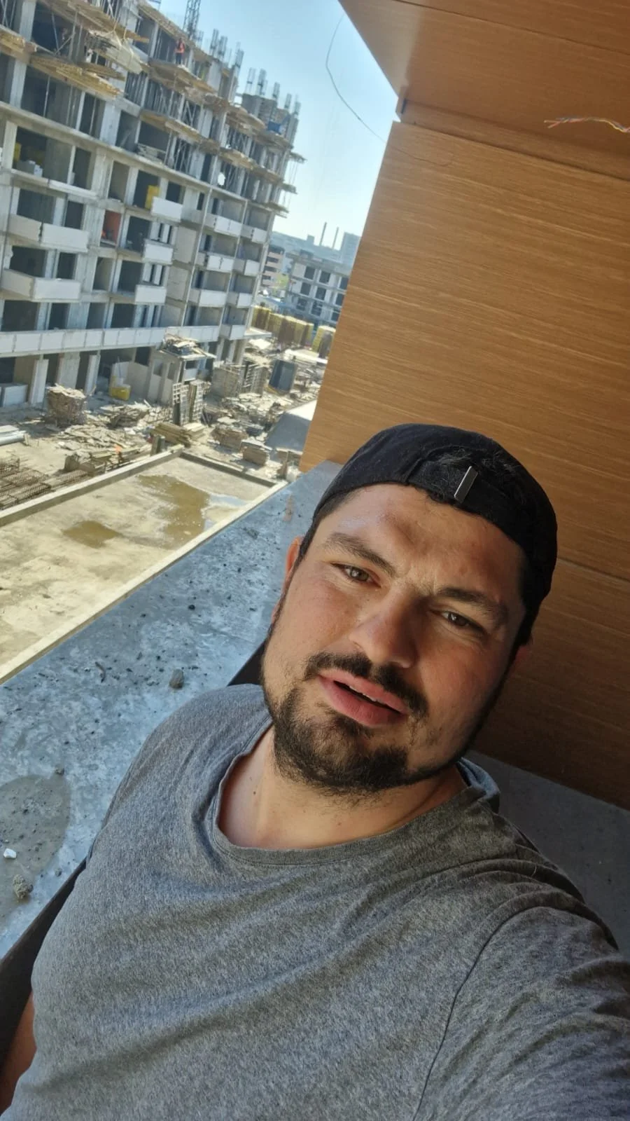 A man taking a selfie on a balcony with a construction site and partially built building in the background.