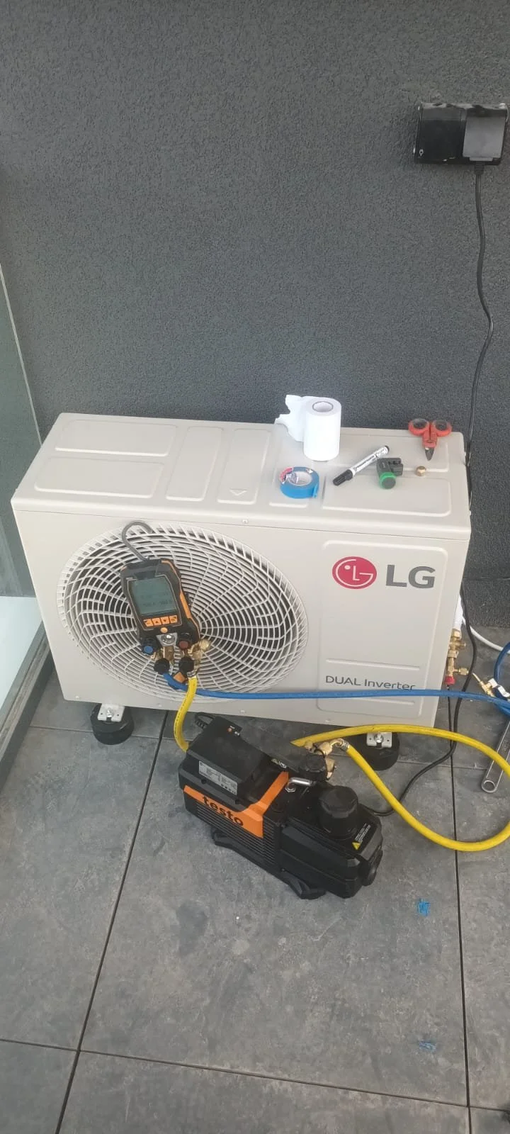 HVAC technician's work area with a LG dual inverter air conditioning unit, tools, and equipment, including a digital manifold gauge, tape, scissors, a flashlight, and a vacuum pump, on a tiled floor.