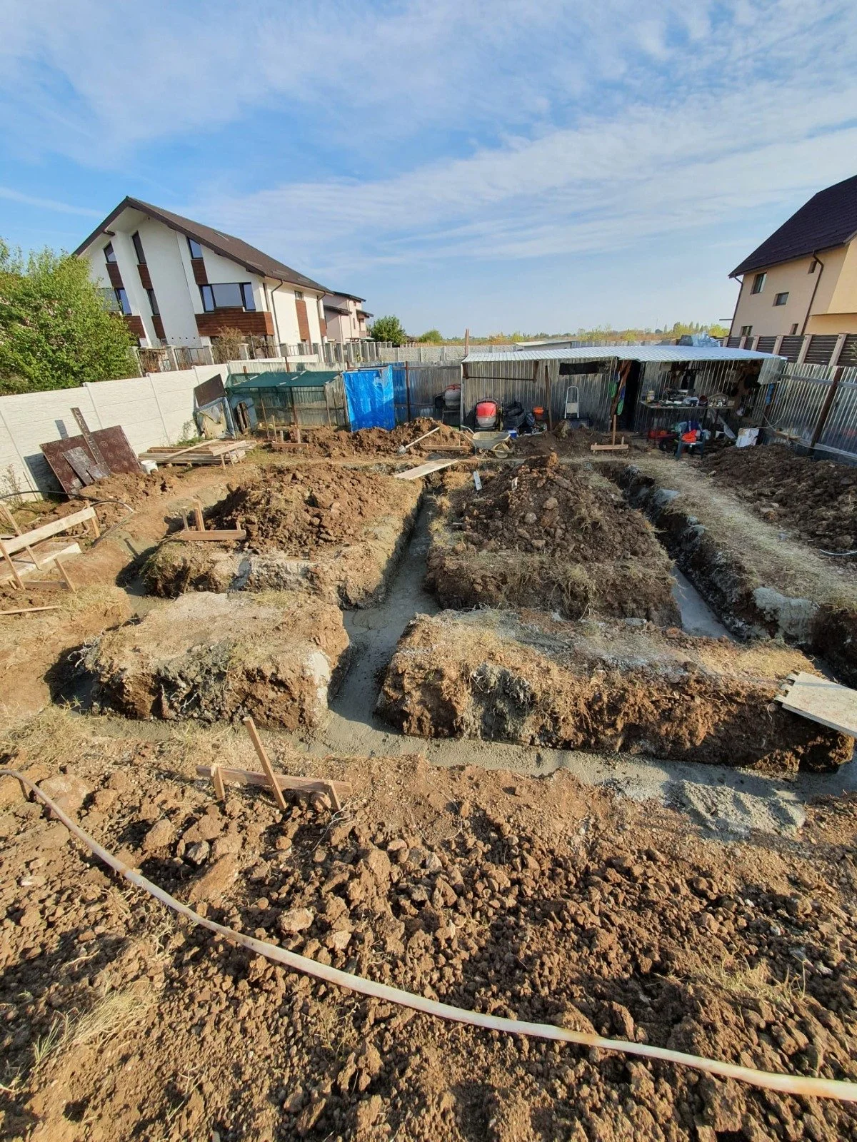 Construction site with trenches and piles of dirt, surrounded by residential houses and fencing, under a partly cloudy sky.