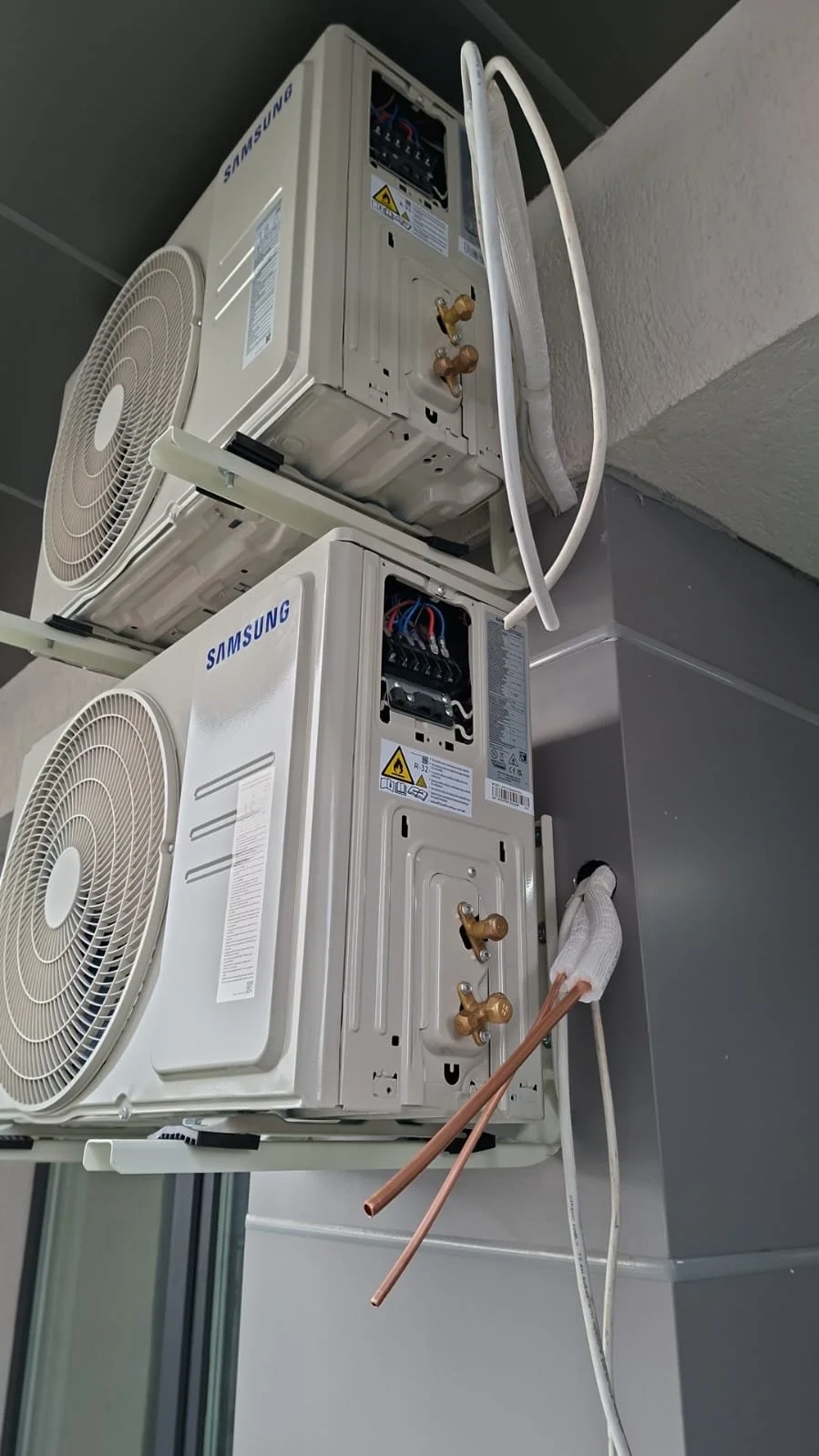 Two wall-mounted Samsung air conditioning units installed in a building, with their covers removed showing internal components, with copper pipes and electrical wiring connected.