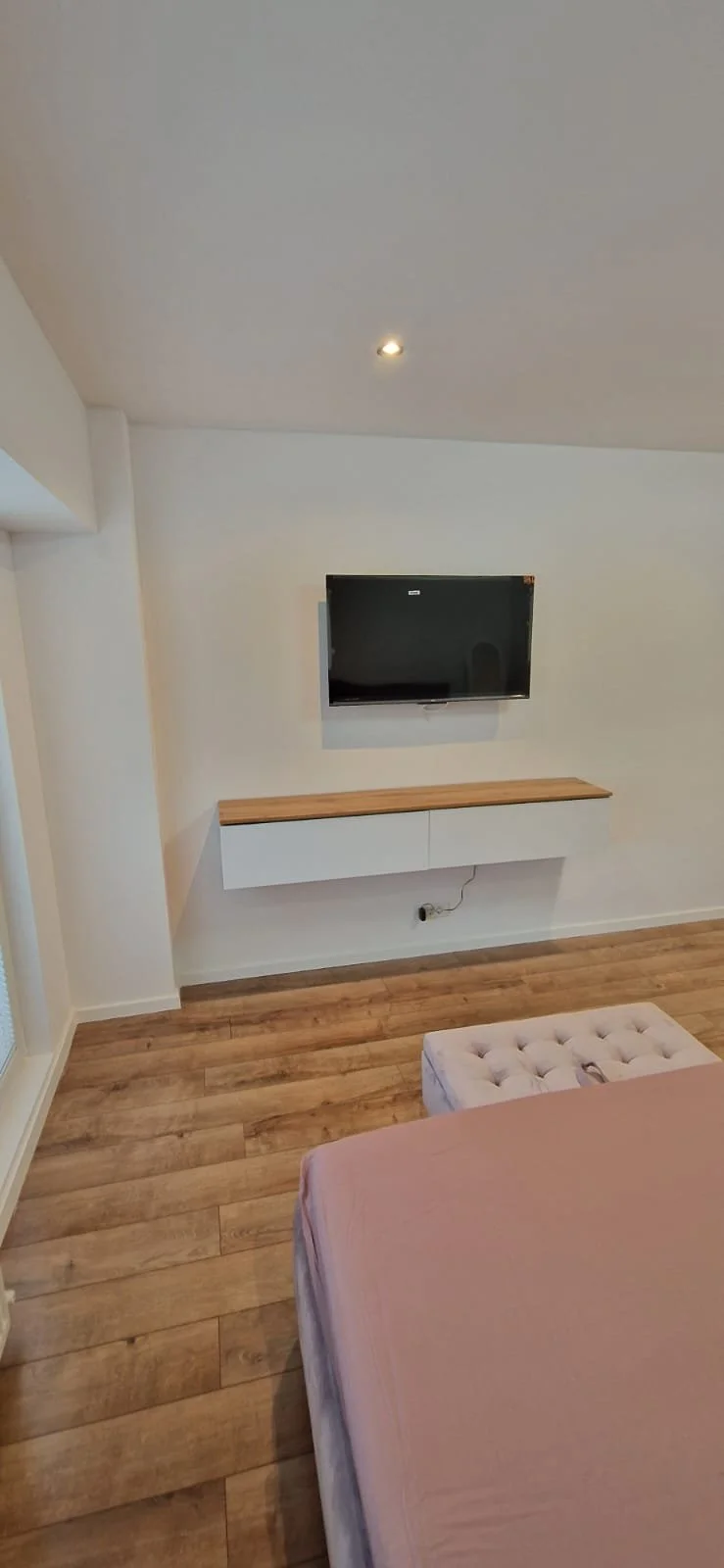 A minimalist living room corner with a mounted flat-screen TV, a white floating cabinet with a wooden top, a tufted ottoman, and a bed with pink bedding on a hardwood floor.
