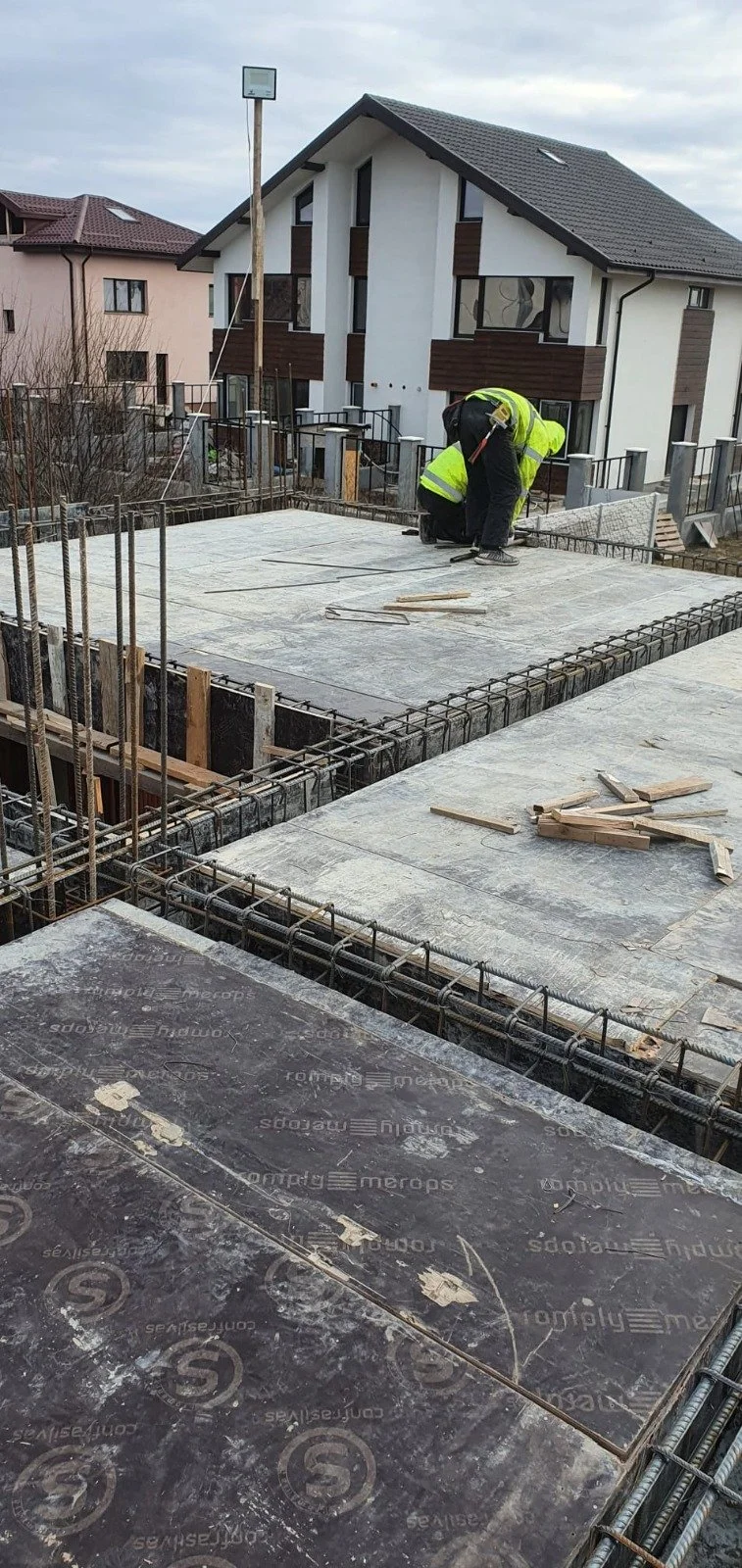Construction worker in a neon yellow safety jacket and black pants kneeling on a concrete building slab at a construction site, surrounded by rebar and construction materials, with residential houses in the background.