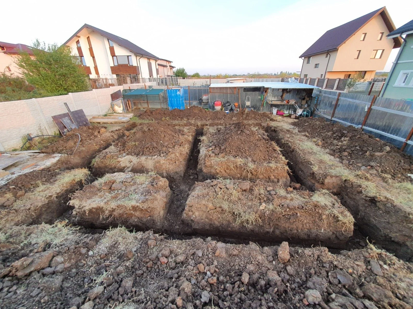 A backyard with several dug trenches for landscaping or construction, surrounded by a fence, with houses and garden tools in the background.
