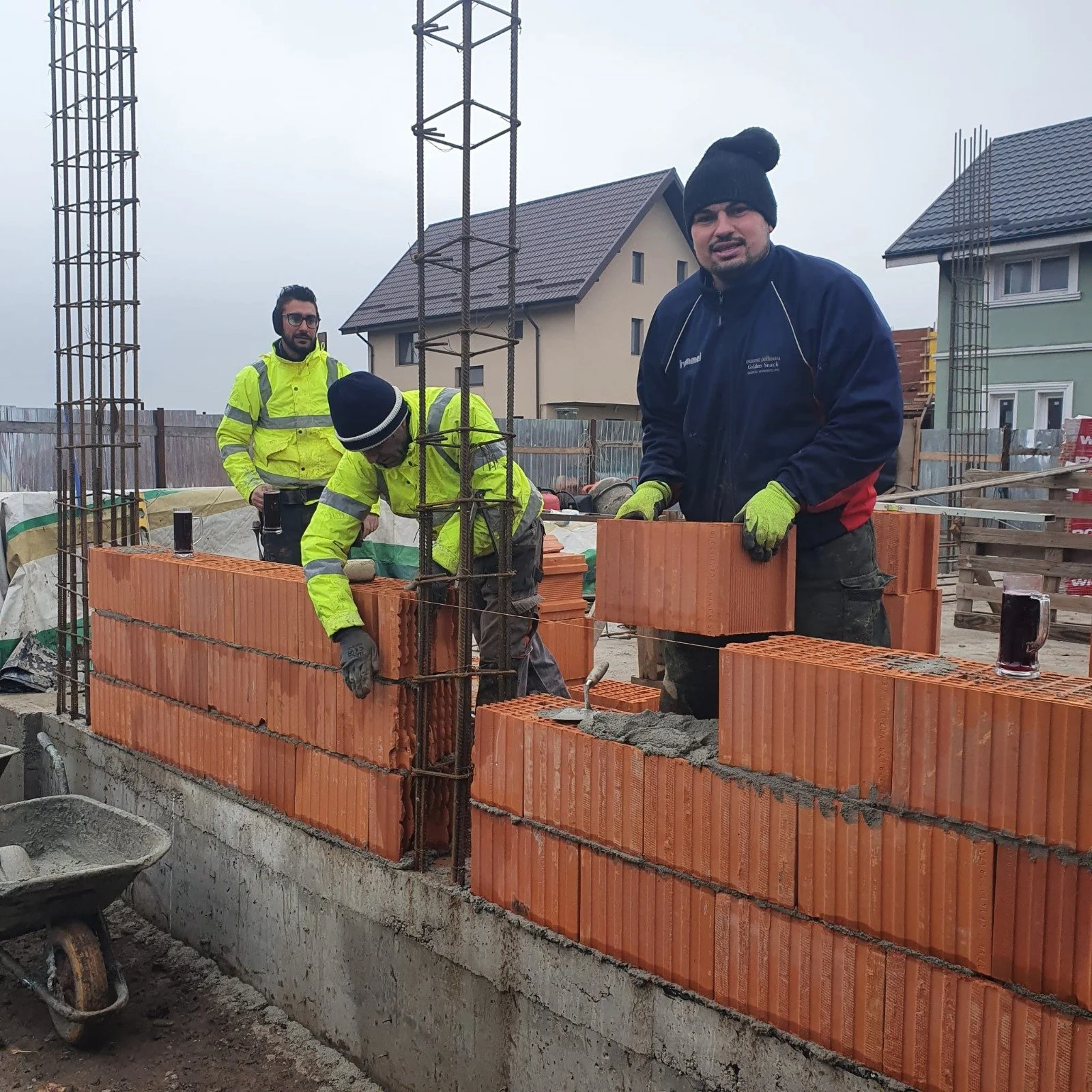 Construction workers building a brick wall on a construction site, with a partially built brick wall and steel rebar in the foreground, and houses in the background.