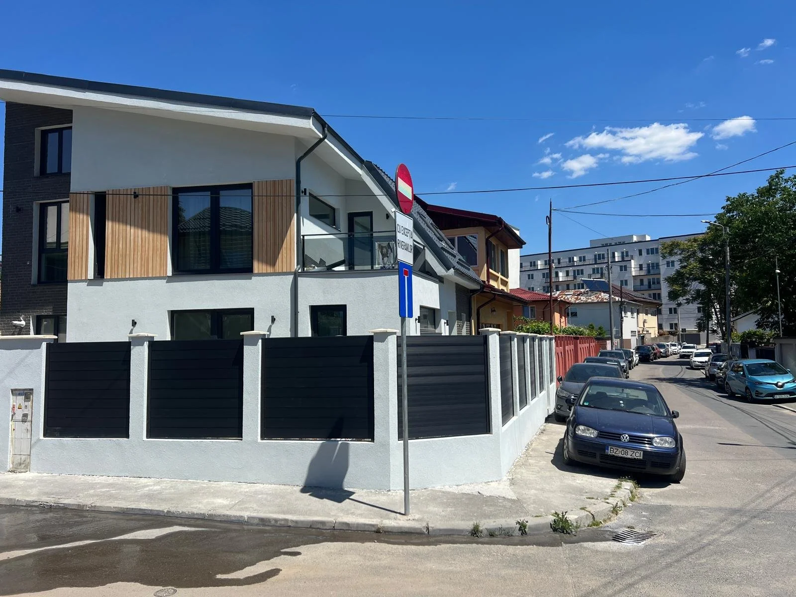 A residential street with modern and traditional houses, parked cars, a clear blue sky, and a street sign indicating no entry and parking restrictions.