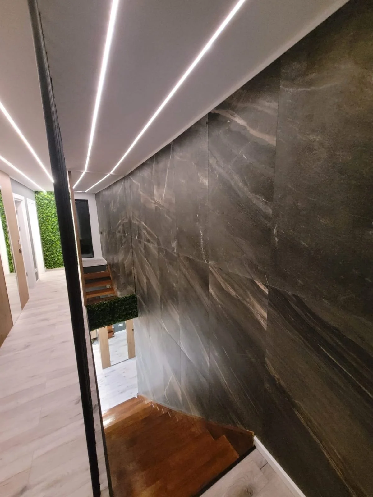 Interior view of a modern staircase with dark marble wall, wooden steps, and LED strip lighting on ceiling.