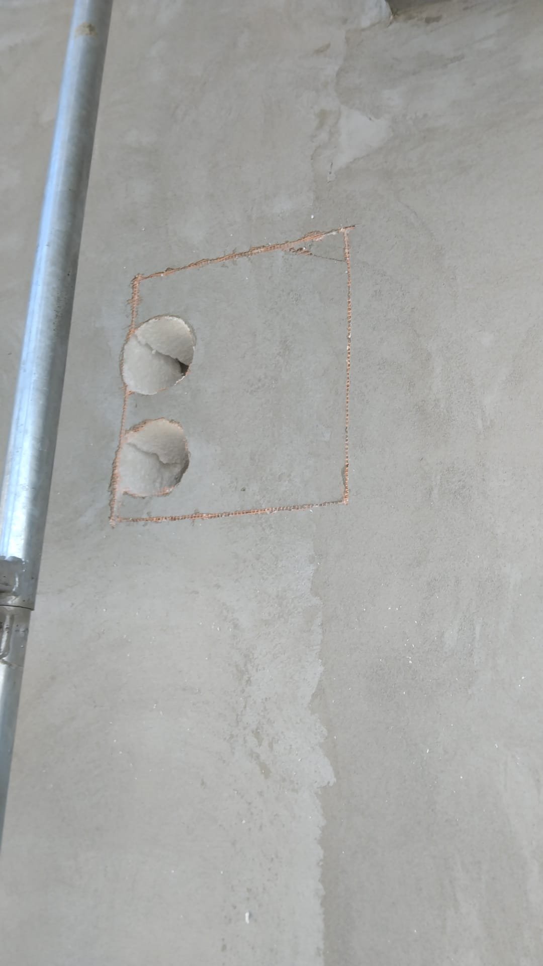 A section of a concrete floor with a rectangular outline and two circular holes drilled inside it, partially covered by a metal scaffold.