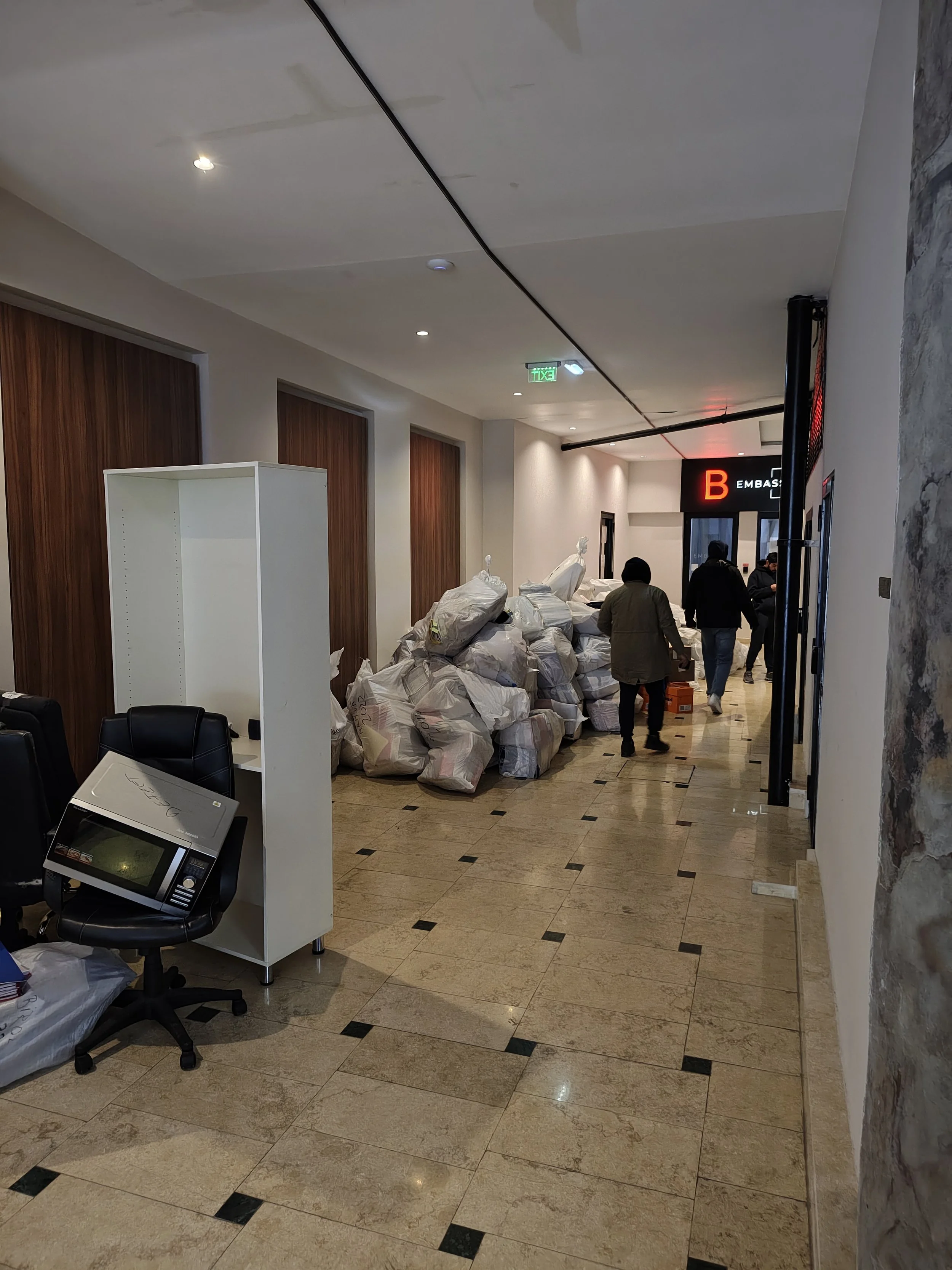 Indoor hallway with piles of plastic bags, possibly filled with trash or materials, along the corridor. Several people walking away from the camera. The hallway has a tiled floor, wooden wall panels, and a ceiling with recessed lighting. There is a white shelving unit and a black office chair with a monitor.