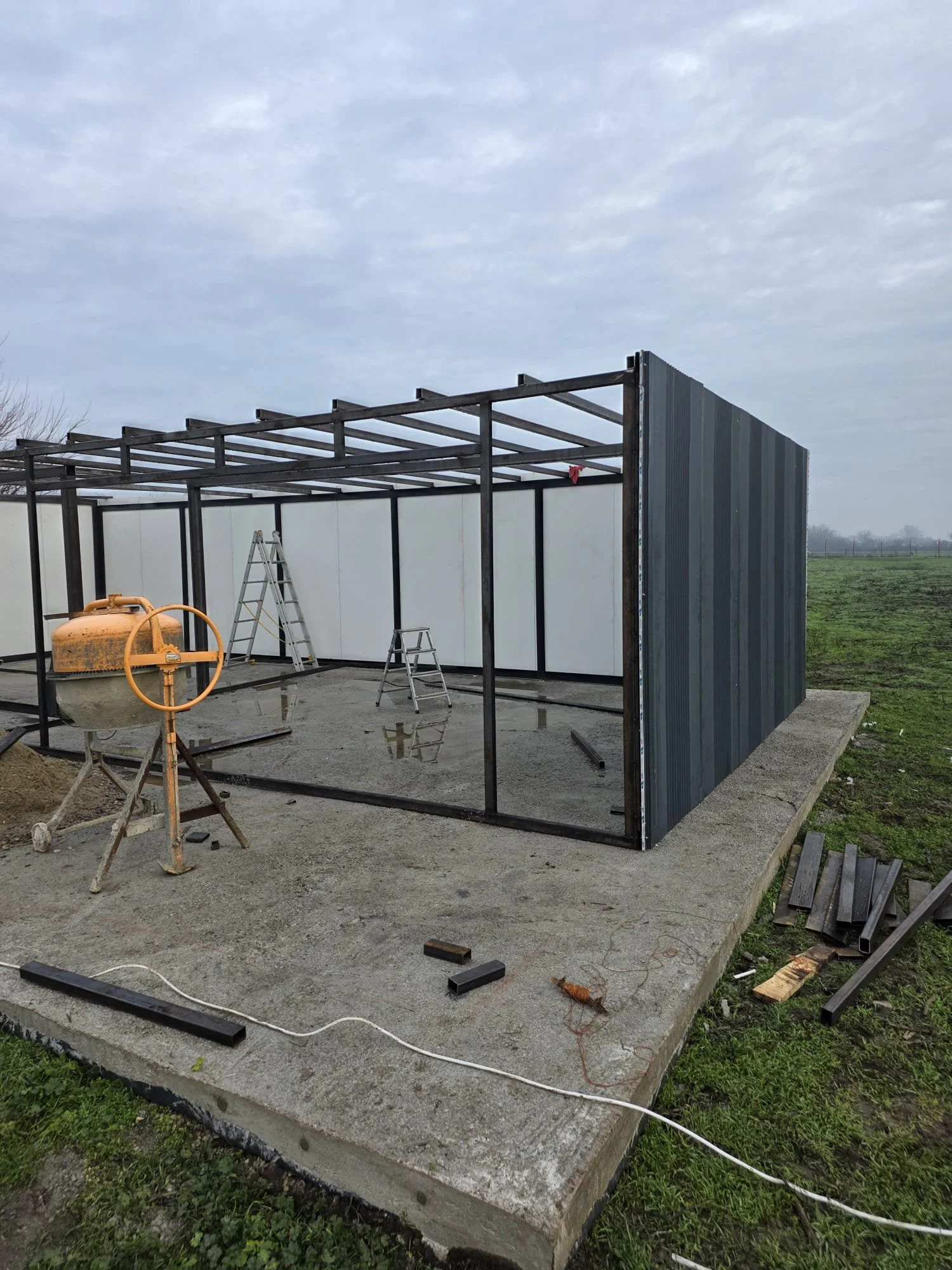 Construction site with a metal-framed structure on a concrete foundation, construction tools, a lime-green concrete mixer, and ladders, in an open field under a cloudy sky.