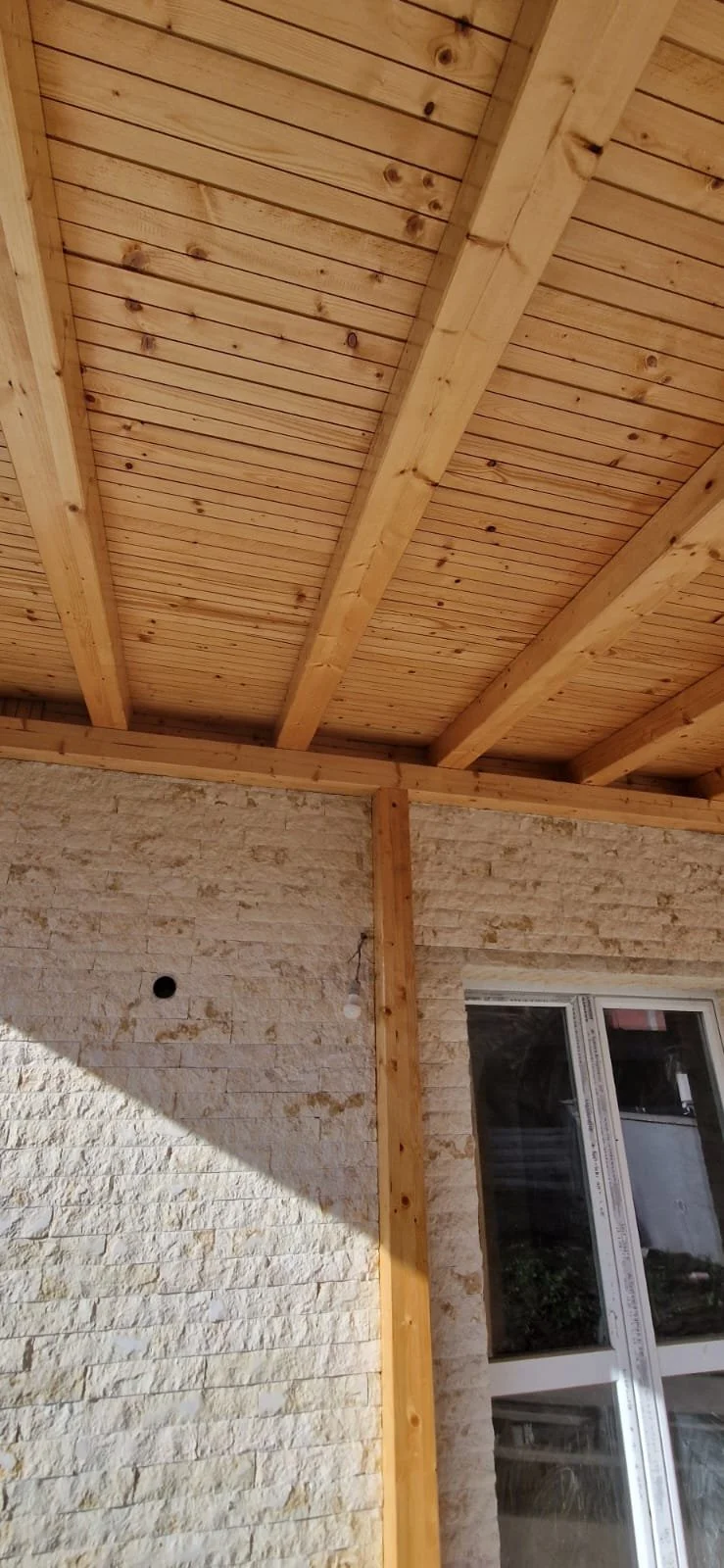 Interior view of a room under construction with a wooden ceiling, exposed beams, and a stone wall with a window.