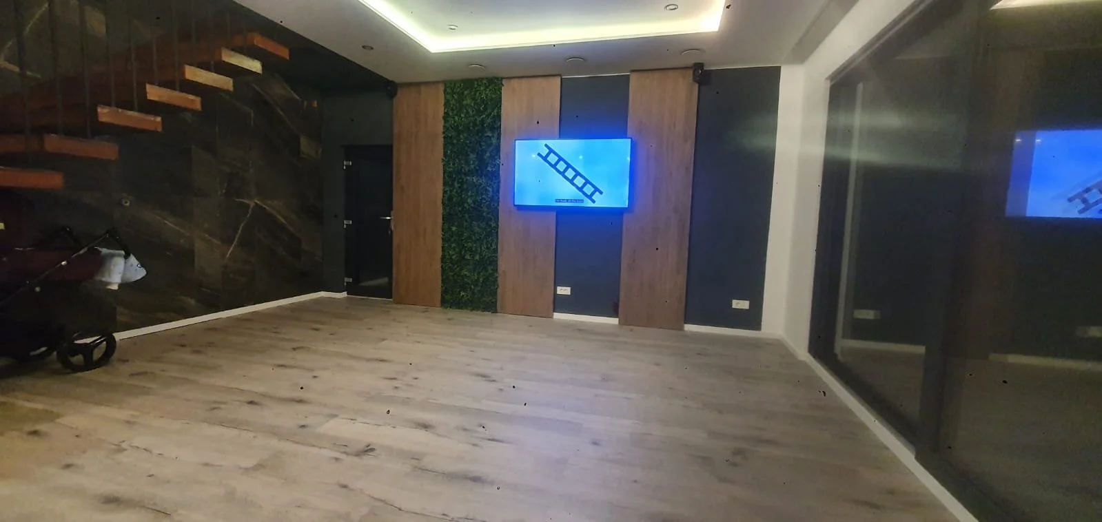 Empty room with wooden flooring, a staircase on the left, a large window or glass door on the right, and a wall-mounted screen displaying a ladder icon in the center of the back wall.