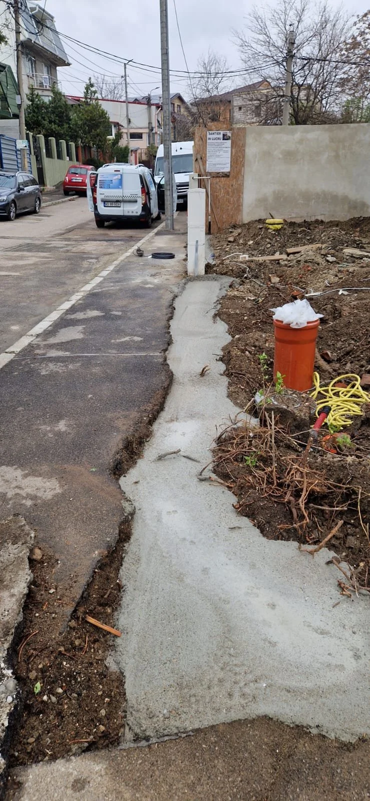 Construction work on sidewalk with fresh concrete, construction materials, and parked cars in the background.