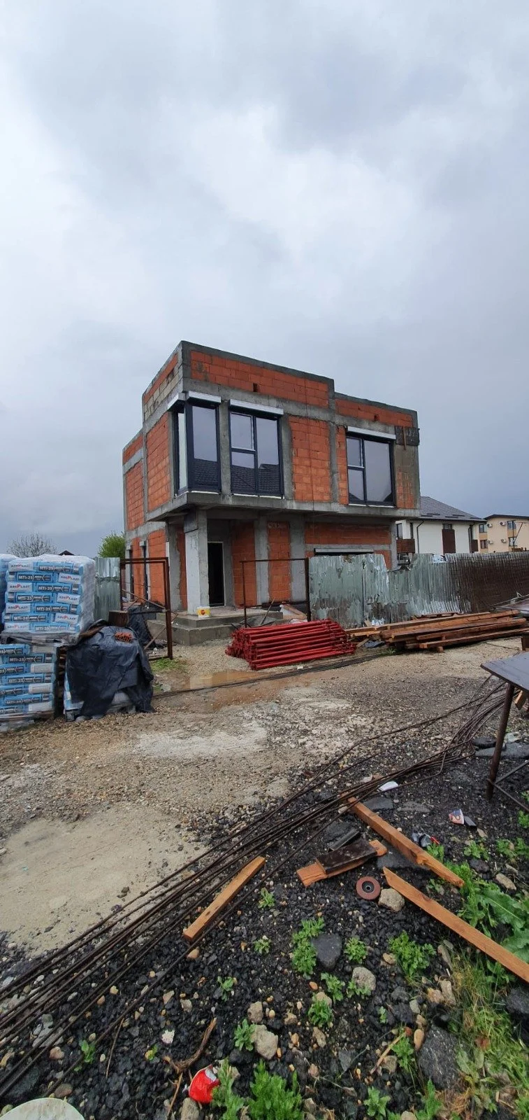 Under construction modern two-story brick house with large windows, construction materials, and muddy ground in the foreground under cloudy sky.