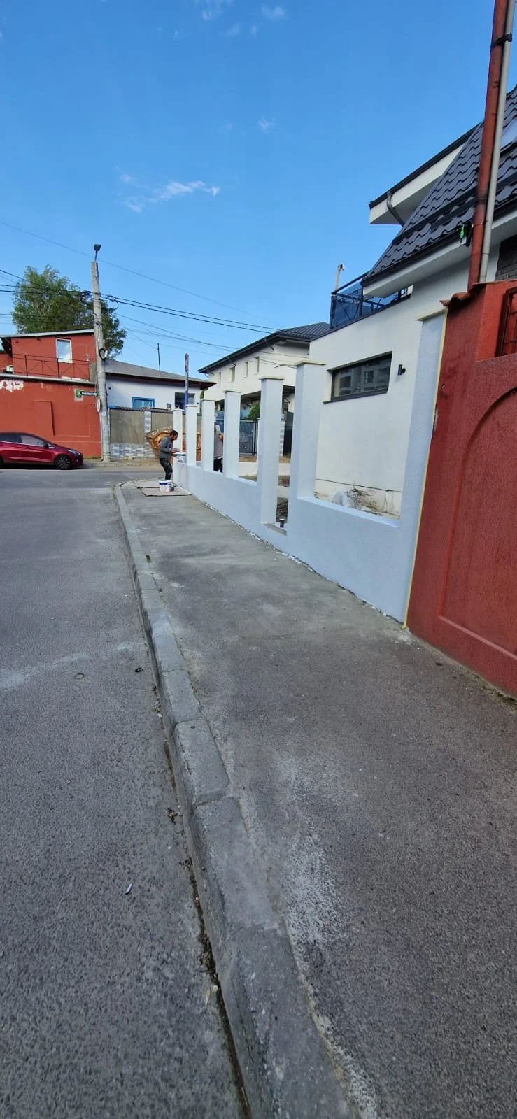 A white half-wall with vertical slits under construction on a sidewalk next to a red and white house, with a person working nearby. A car is parked on the street, and a red building is visible in the background under a blue sky.