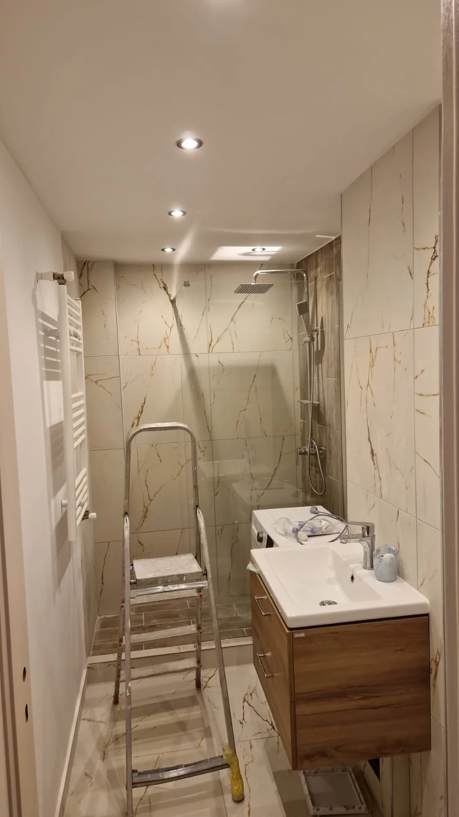 Bathroom with marble wall tiles, a shower area with a rainfall showerhead, a small sink vanity, a ladder, and a towel warmer.
