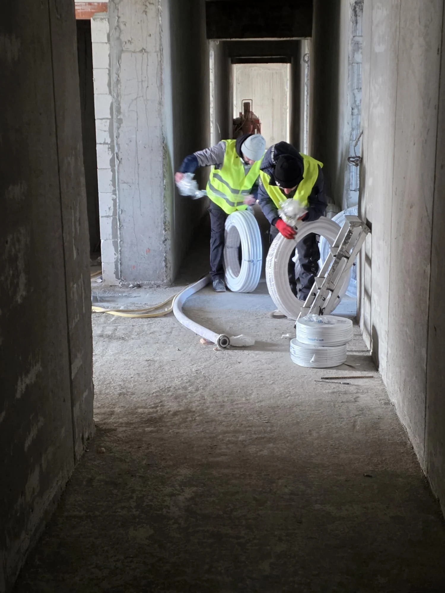 Two construction workers wearing yellow safety vests and gloves working with white coils of electrical wire in a building under construction.