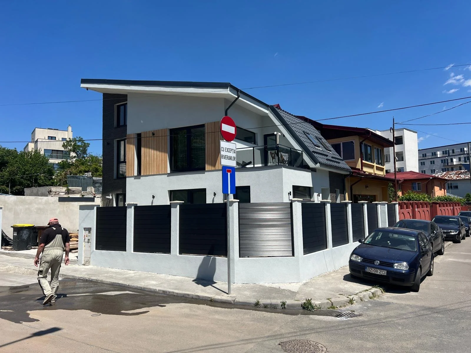 Modern two-story house with white and dark gray exterior, wooden accents, and large windows, surrounded by a black fence. Several parked cars and a man walking past on the street. Clear blue sky.