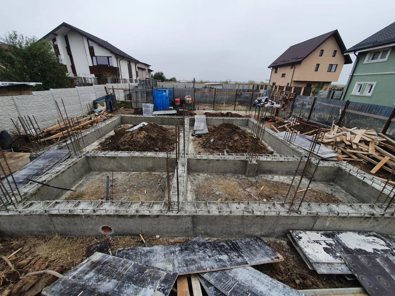 Construction site with concrete foundation and rebar for a new building, surrounded by construction materials and houses in the background.