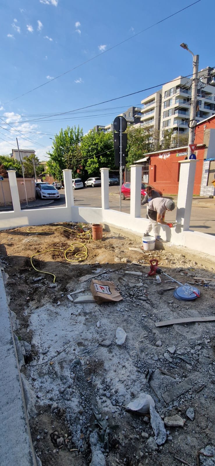Two workers are constructing a white wall on a construction site with dirt and debris in the foreground. There are several cars parked and a multi-story apartment building in the background, along with a clear blue sky.