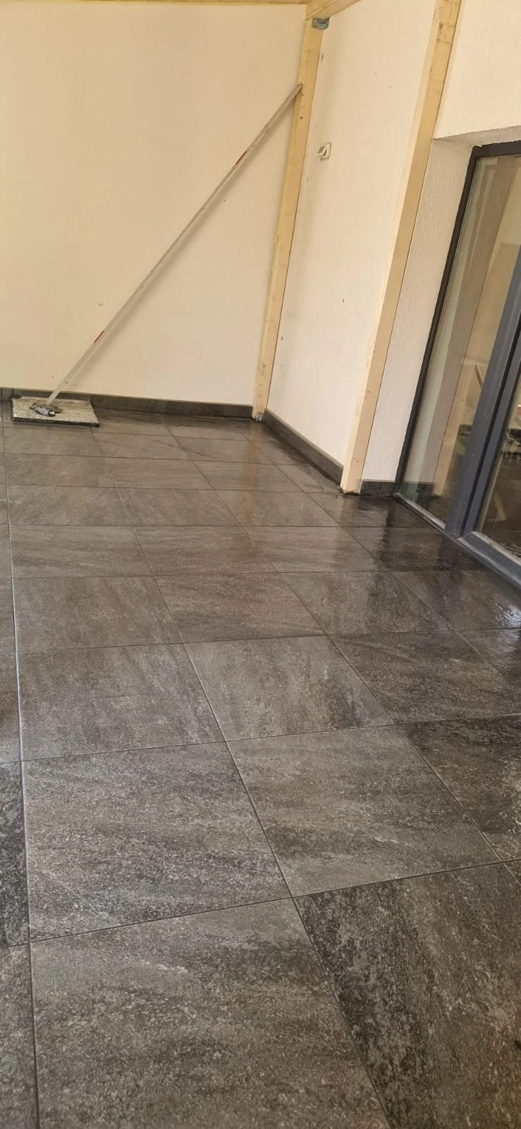 Interior of a room with newly installed gray ceramic tile flooring, a partially framed wall, and a window or glass door.