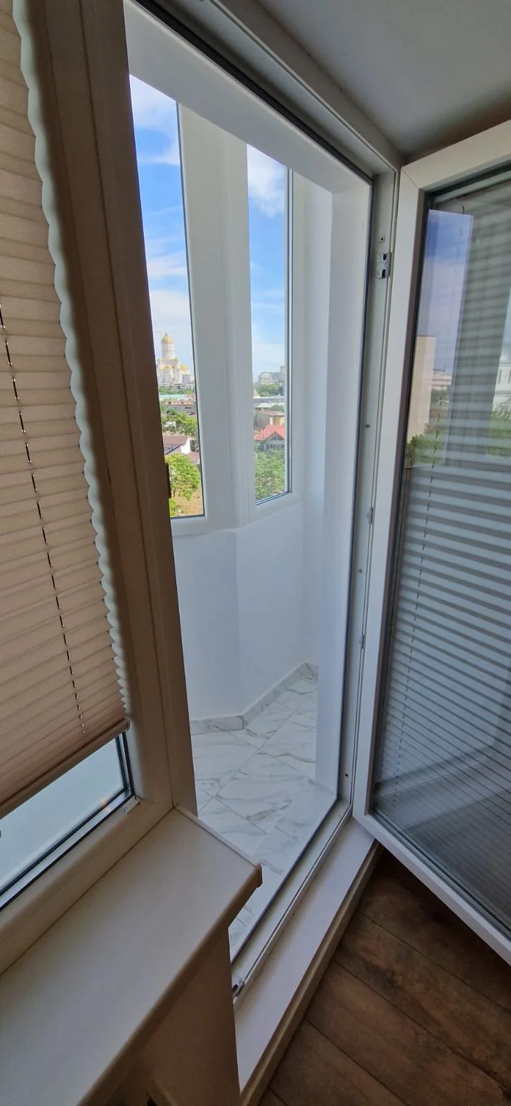View through a glass balcony door showing a cityscape with a tall gold-domed building and lush green trees, with a partly cloudy sky.
