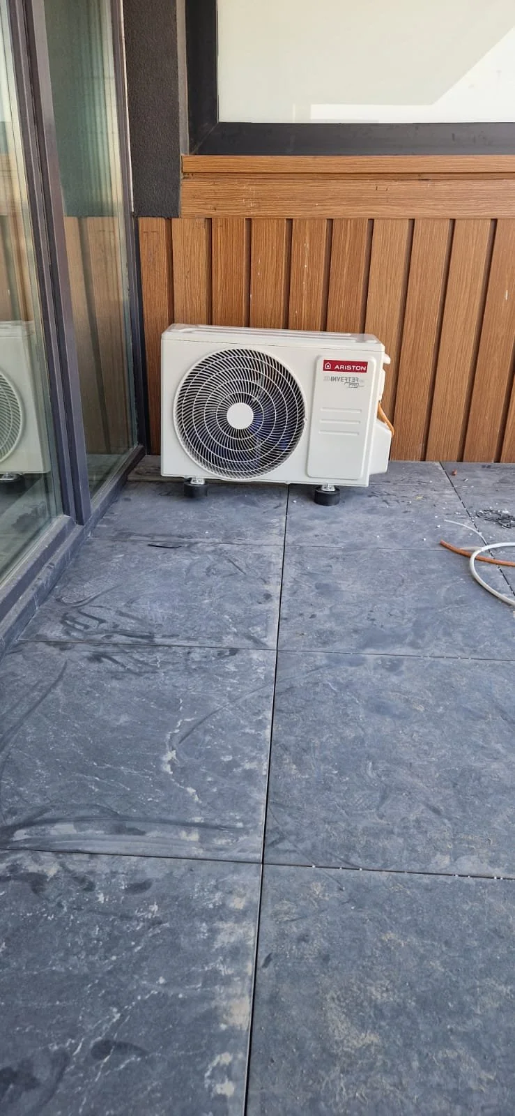 Outdoor air conditioning unit on a stone patio near a building with wood siding and a glass sliding door.