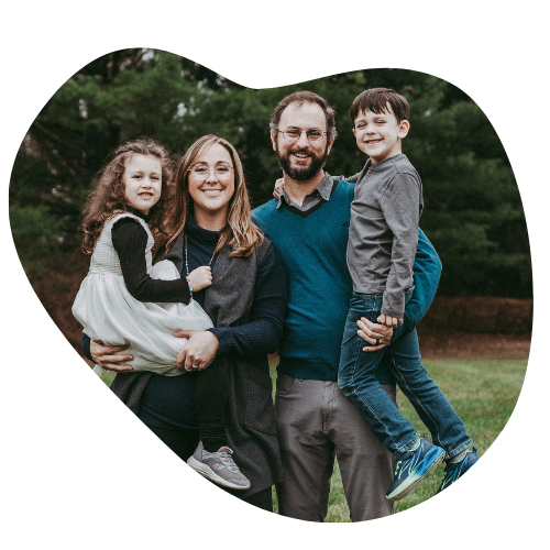 A family of four standing outdoors on a grassy area with trees in the background, smiling at the camera. The mother and father are holding their two children, a girl and a boy.