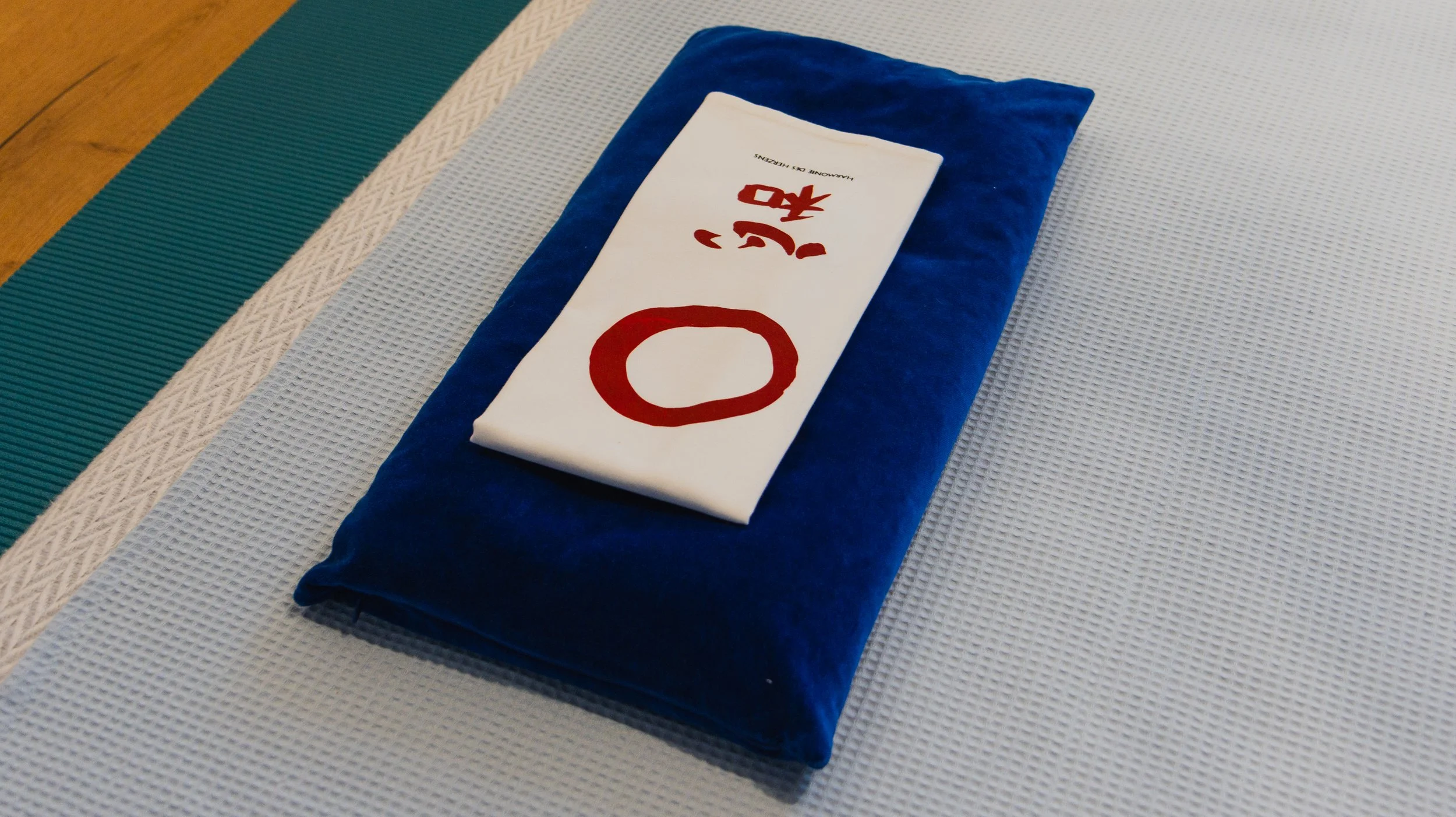 Ein weißes Tuch mit rotem Kreis und japanischen Schriftzeichen liegt auf einem blauen Kissen auf einer hellen Matte.