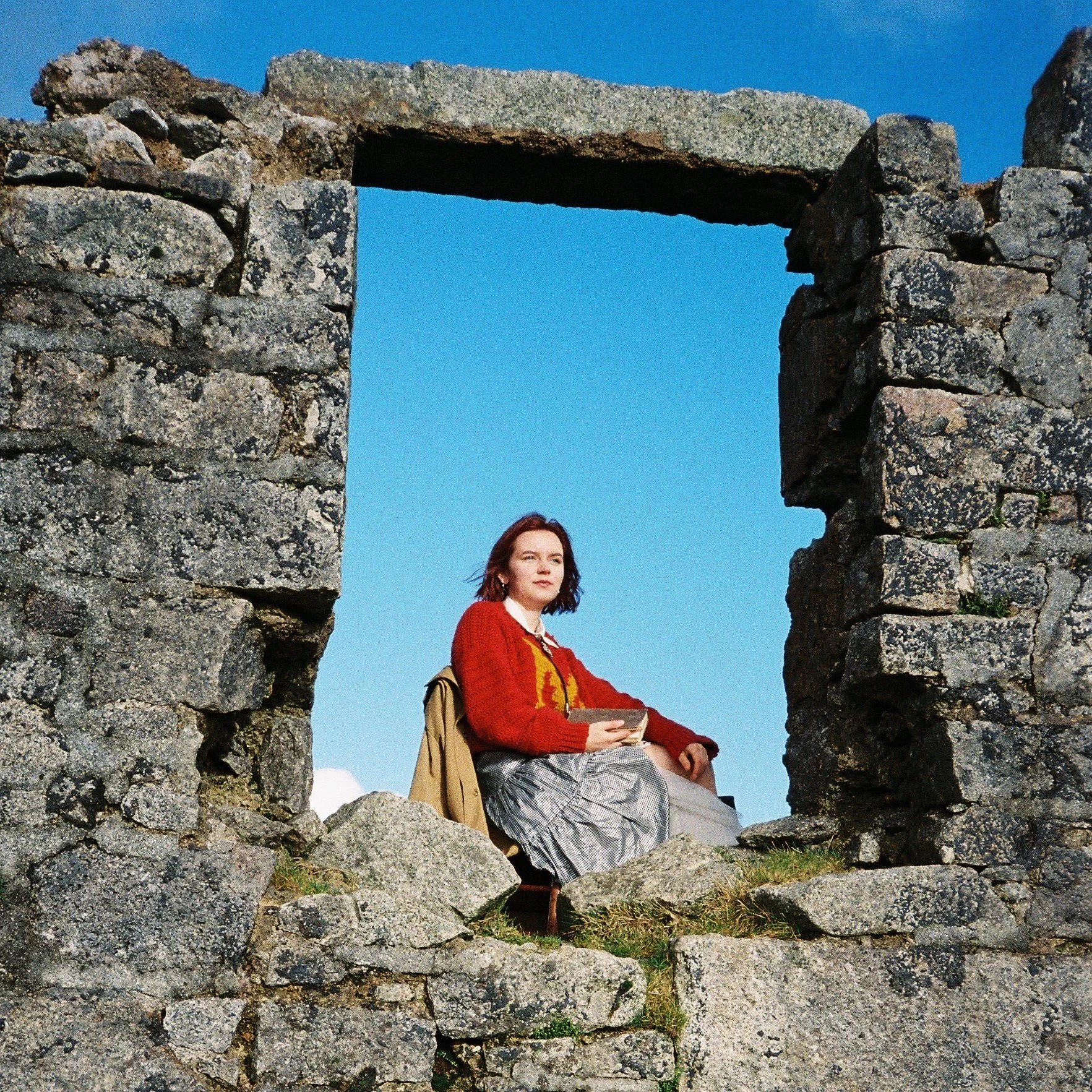 A woman sitting on ruins of a stone window frame, holding a book, with a clear blue sky in the background.