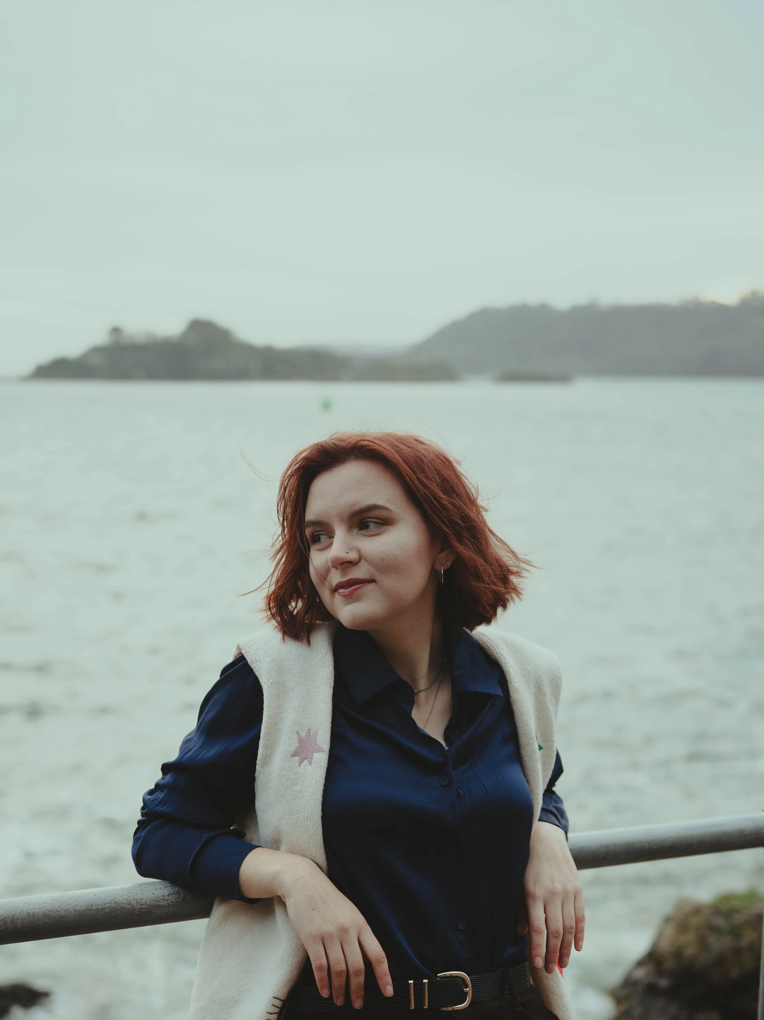 A woman with short red hair standing by a railing near the sea, with an island visible in the background.