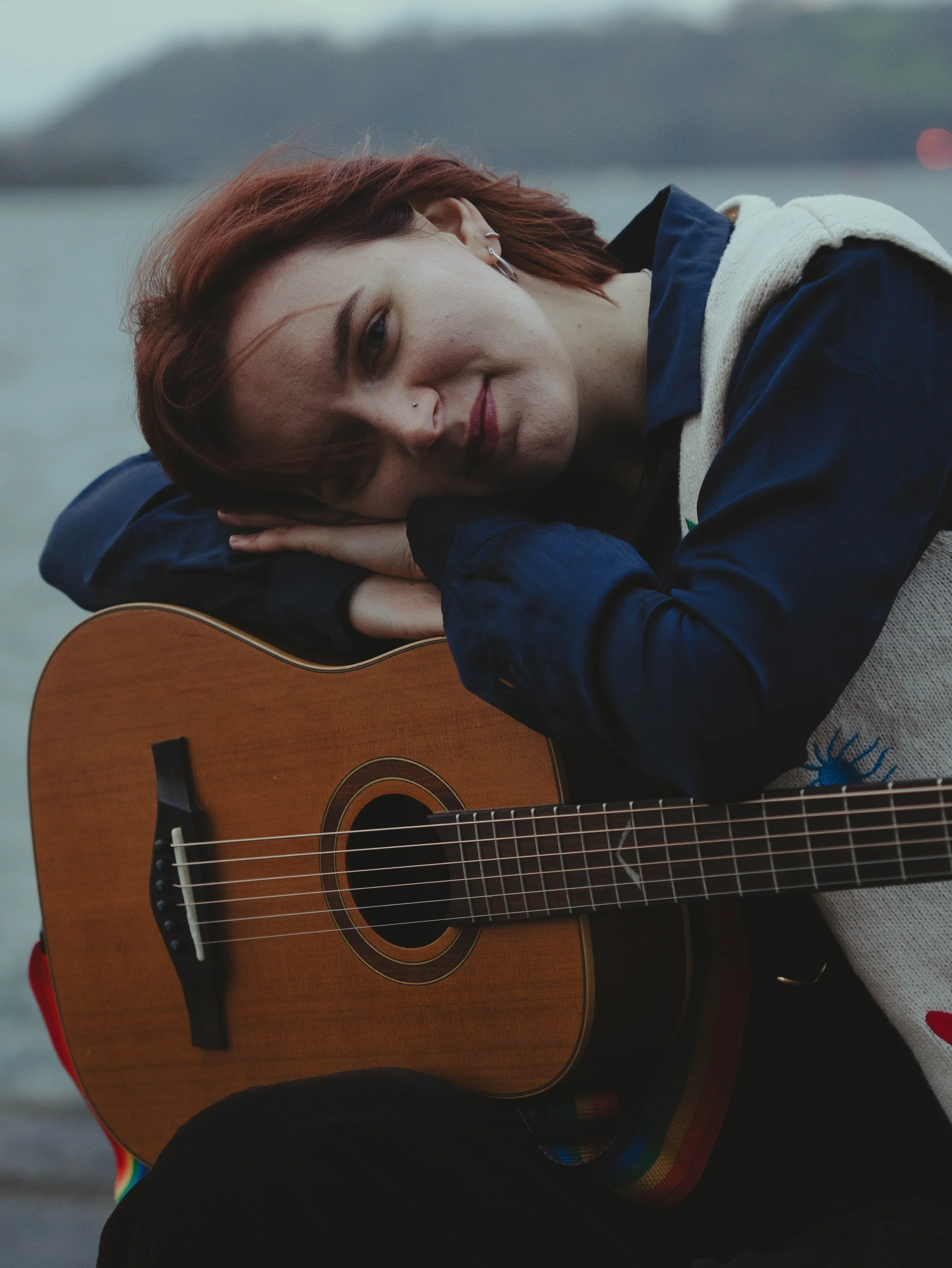 A woman with short reddish hair resting her head on a guitar outdoors, with the sea and distant hills in the background.