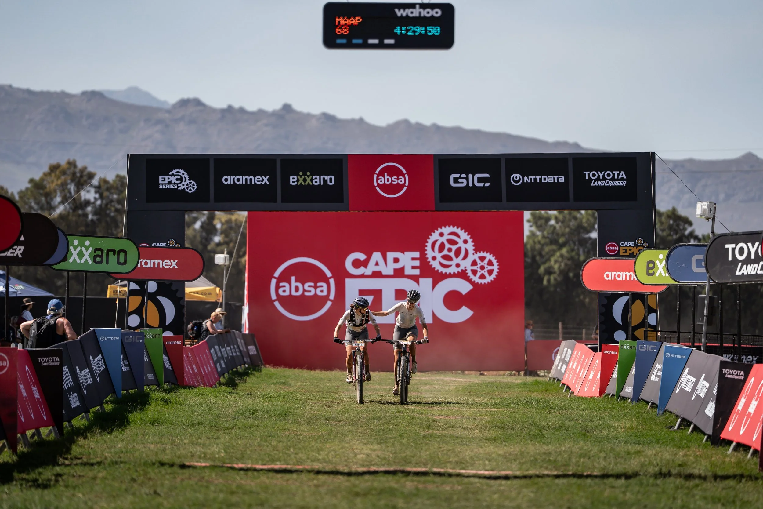 Two cyclists riding bikes side by side toward the finish line at the Cape Epic mountain bike race, with a red backdrop displaying the event logo and a gear symbol, surrounded by sponsor banners and spectators