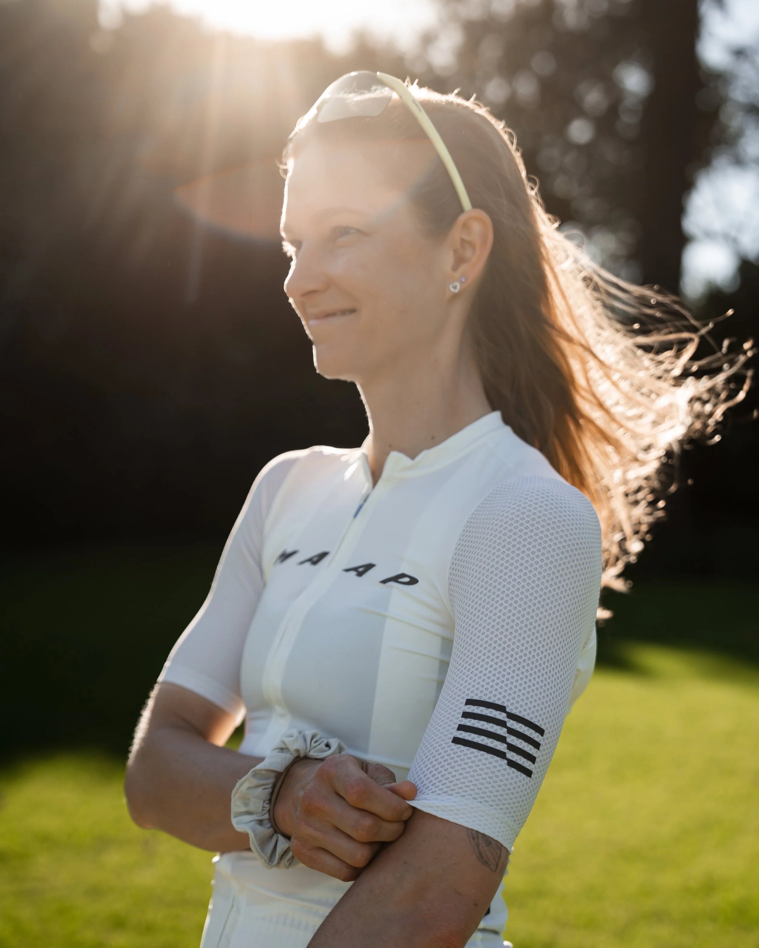 A woman in a white sports jersey standing outdoors in sunlight with arms crossed, smiling and looking to her right.