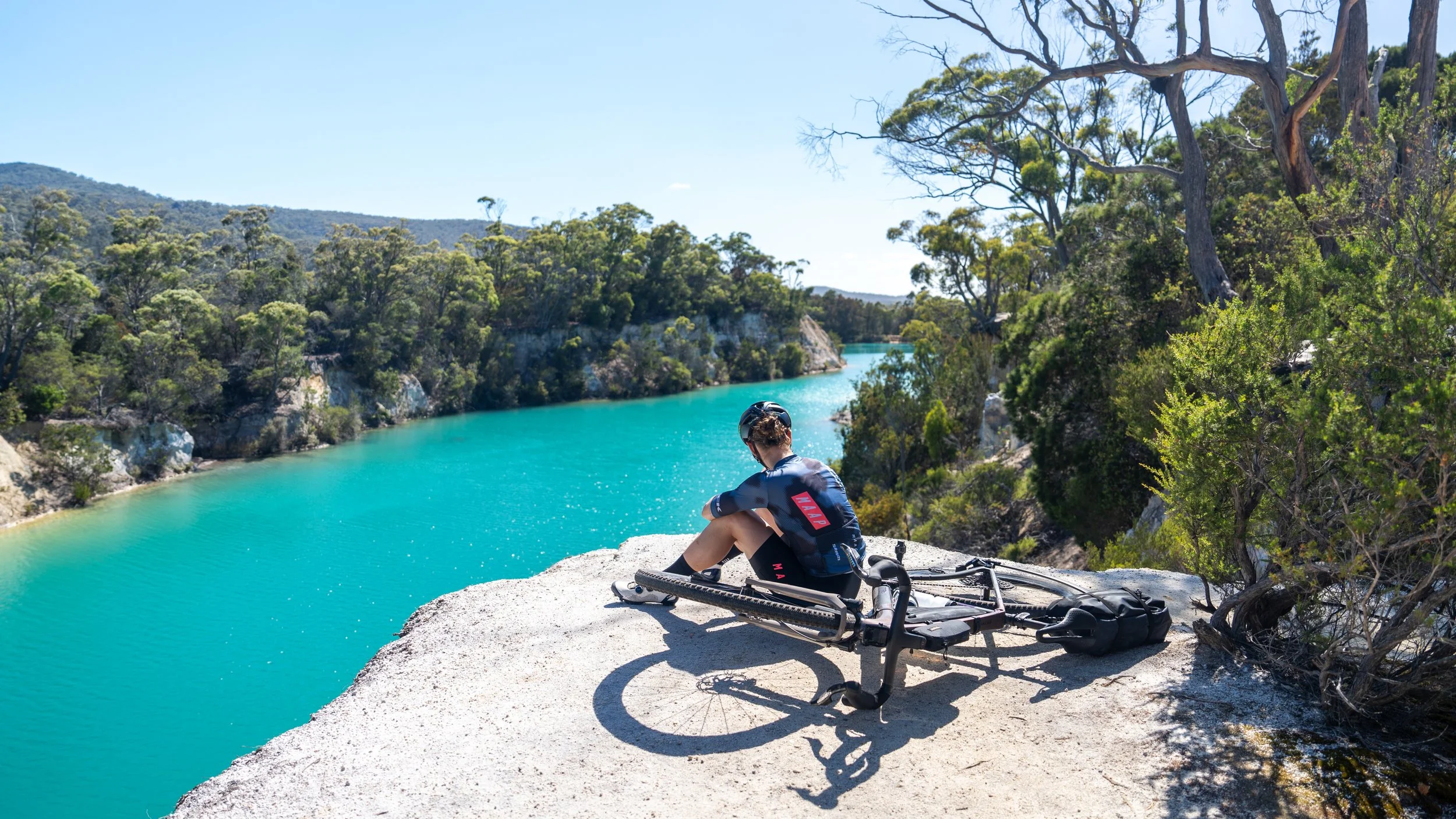 Person sitting on a rocky ledge overlooking a turquoise river with trees and hills in the background, with a bicycle and helmet nearby.