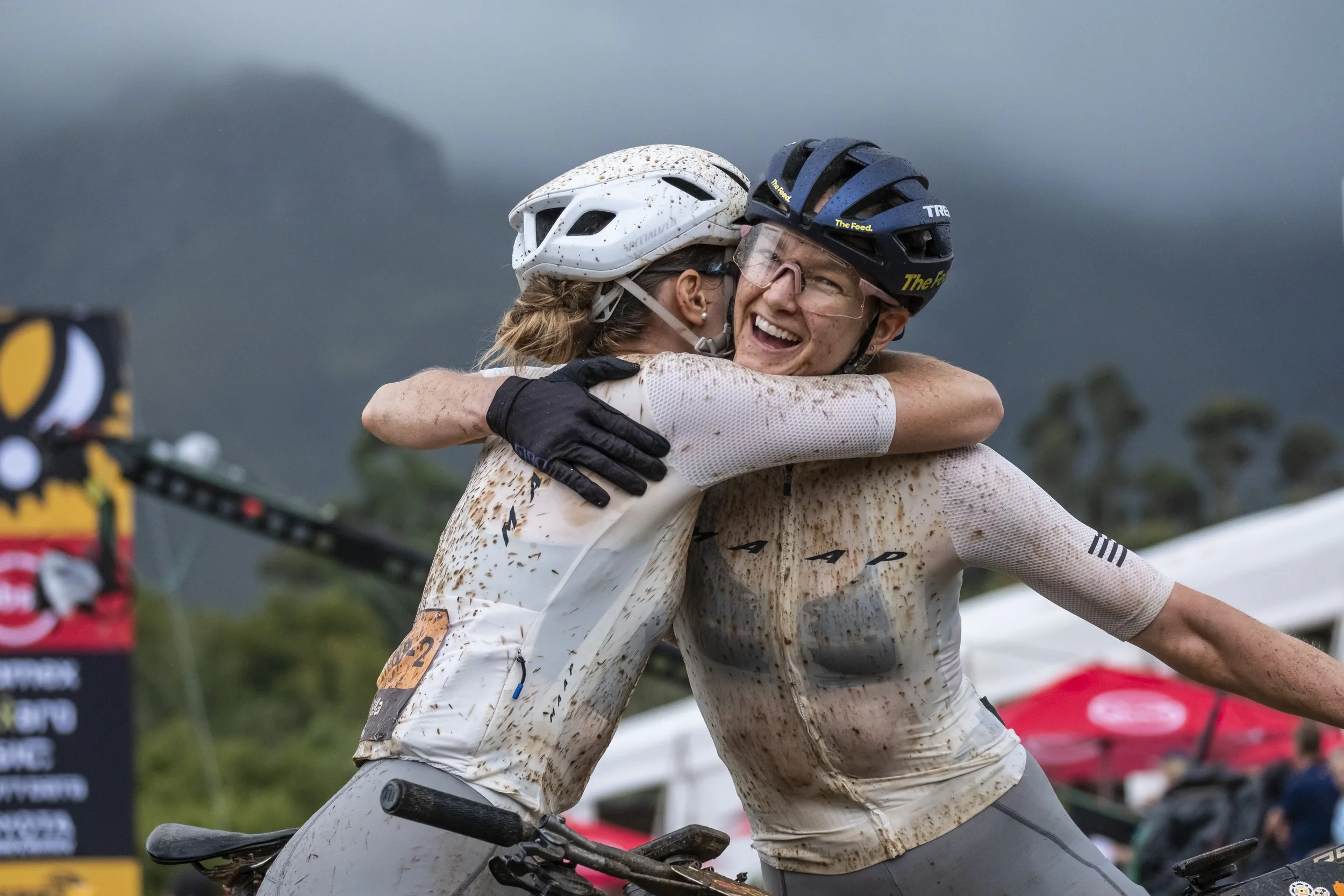 Two cyclists in helmets and muddy jerseys hug and smile after a race, set against a cloudy, mountainous background.