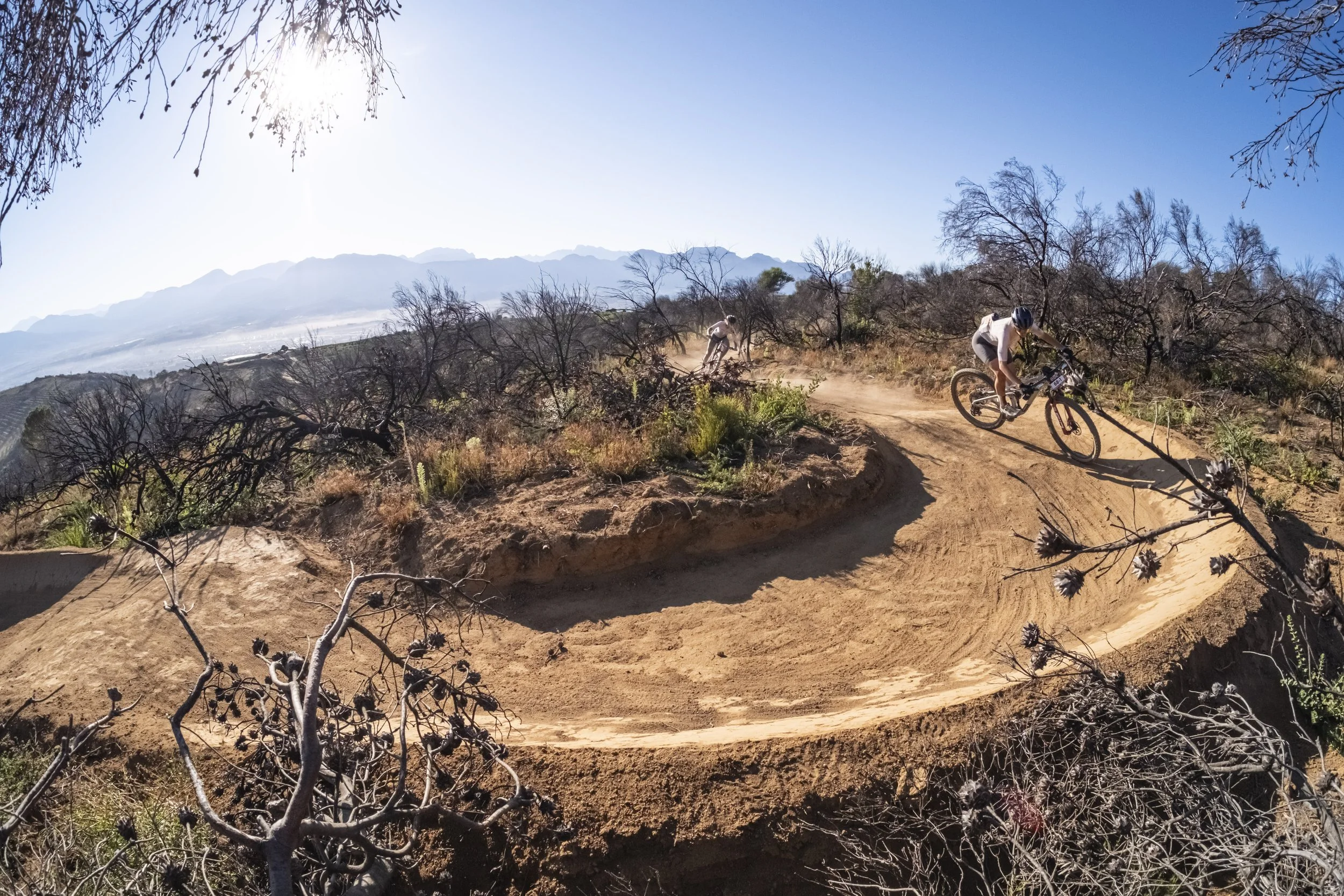 Two mountain bikers riding on a dirt trail in a dry, hilly landscape with sparse bushes and leafless trees, under a clear blue sky.