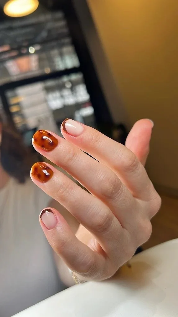 Close-up of a hand with manicured nails, featuring tortoiseshell design on some nails and natural unpolished look on others, in a setting with blurred background.