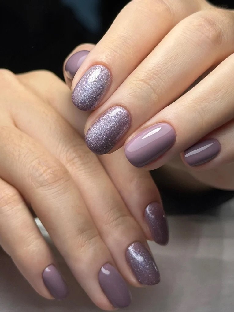 Close-up of hands with manicured nails painted in shades of purple and lavender, some with a shiny finish and others with a glittery or matte texture.