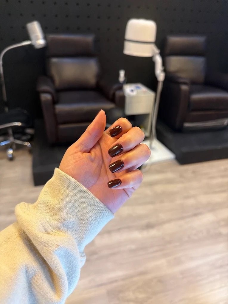 A person's hand with dark brown painted nails, wearing a beige sweatshirt, is shown in the foreground. In the background, there are two brown salon chairs with headrests, a white hair dryer, and a small white table with a hair tool on it. The setting