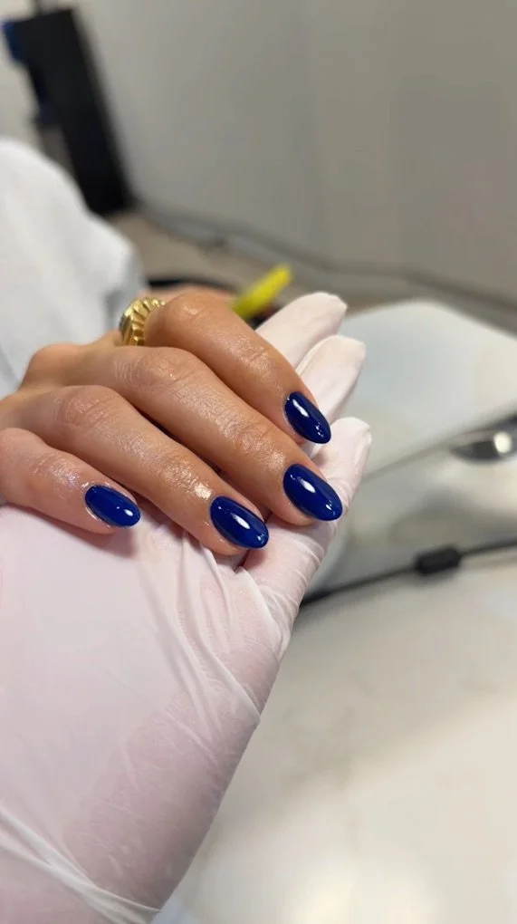 Close-up of a woman with royal blue manicured nails holding a hand with a professional nail technician wearing a white glove. The background shows a salon or clinic setting.