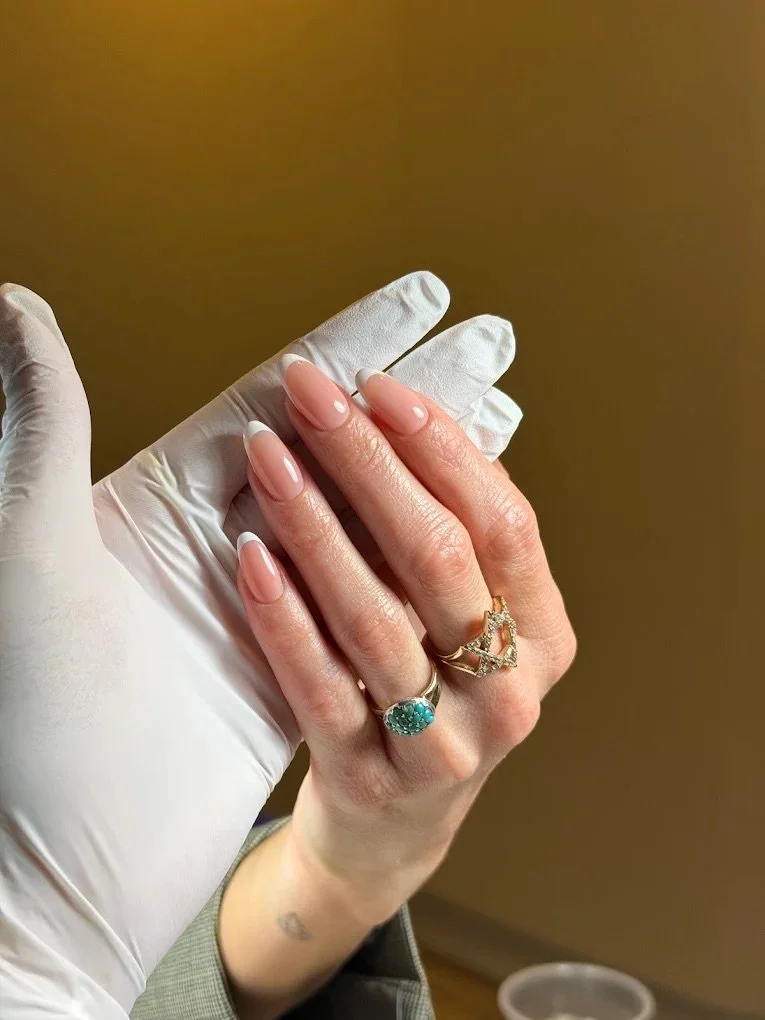 Close-up of a person's hand showing manicured nails and rings, with a gloved finger holding the hand. The rings include an emerald-colored gemstone and a layered gold ring with small diamonds. The background is blurred in a warm tone.