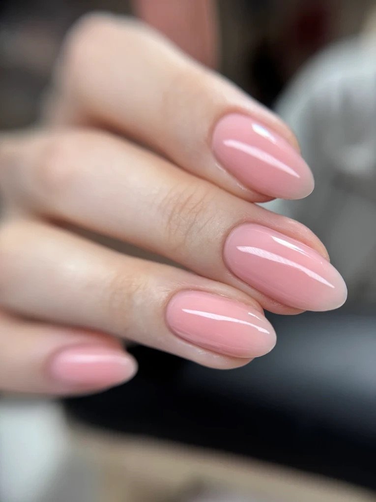 Close-up of a person's manicure with glossy light pink nail polish on neatly shaped oval nails.