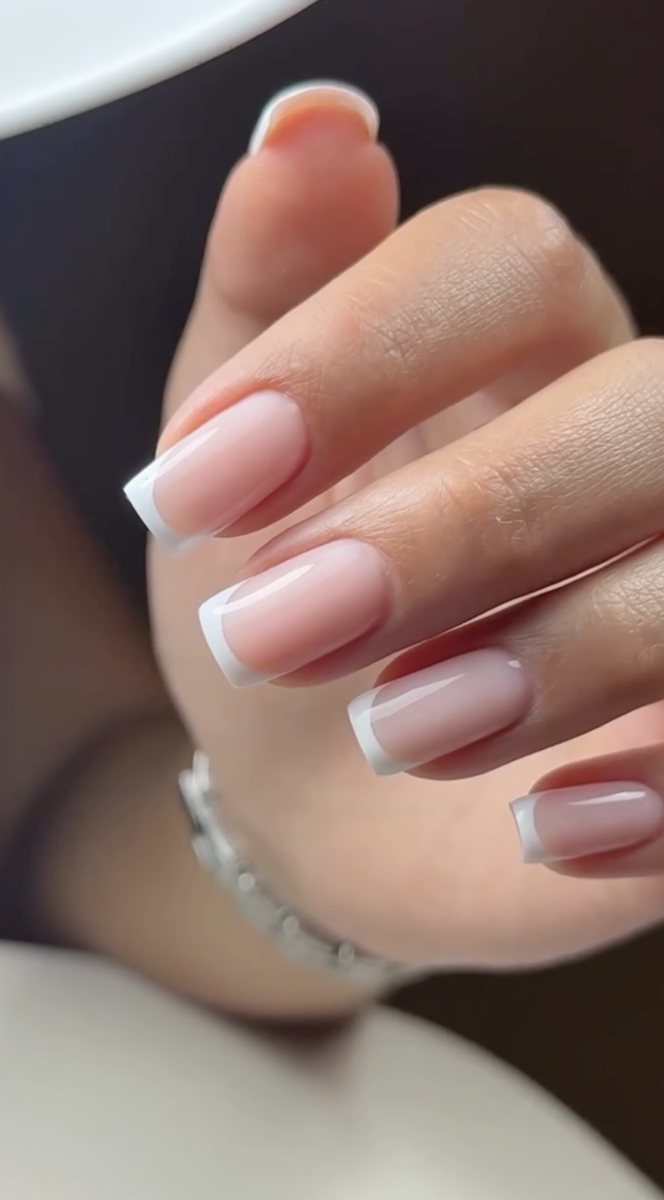 Close-up of a person's hand with well-manicured nails featuring clear, natural-looking nail polish with white tips, and a silver bracelet on the wrist.